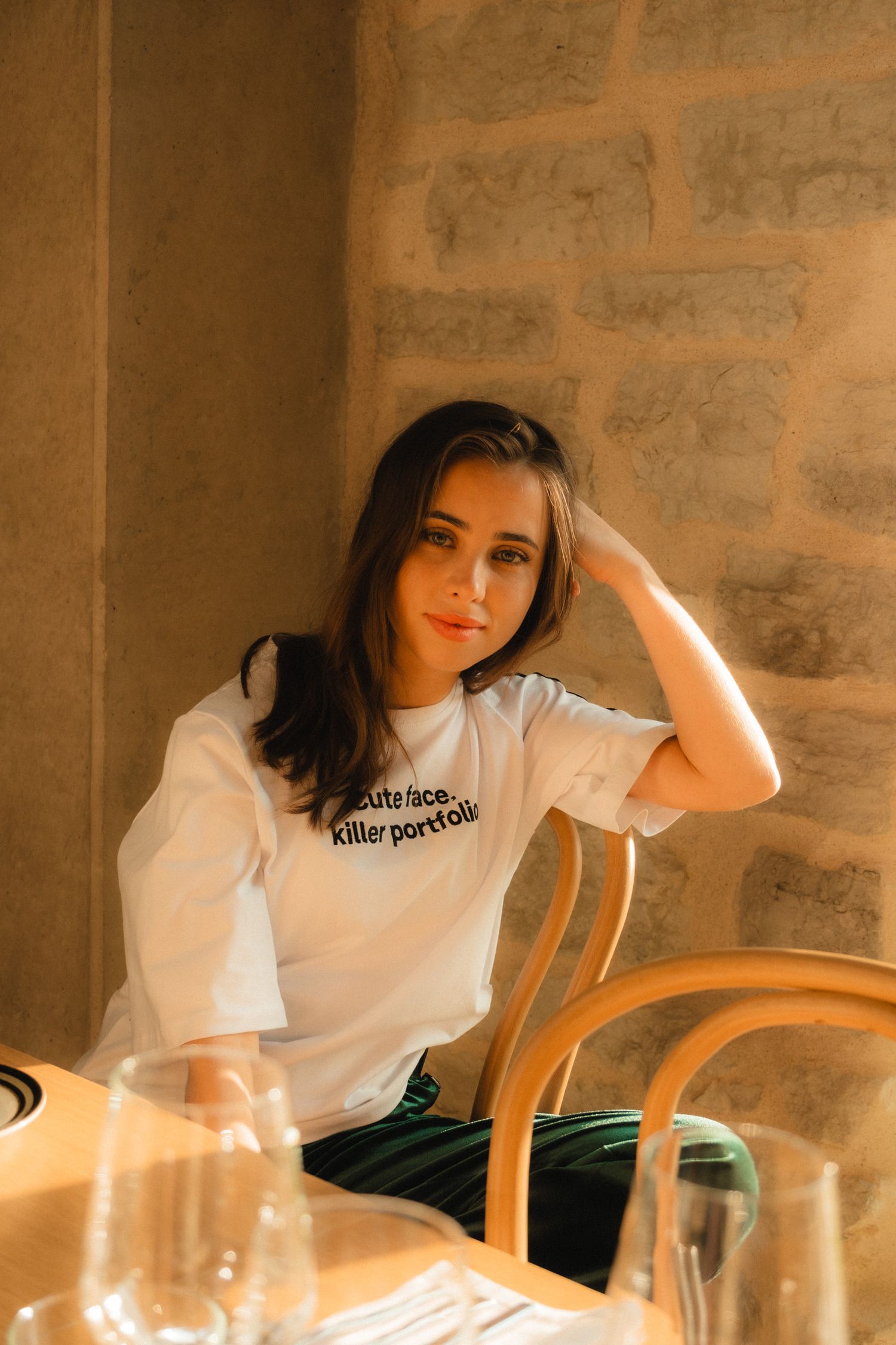 A person in a white t-shirt sits at a table with glassware in warm lighting against a stone wall.