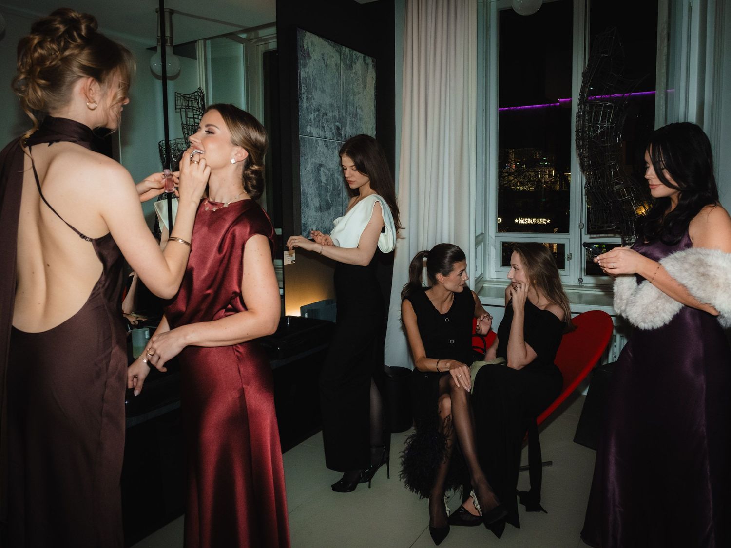 Ladies adjusting make up together in a dim lit bathroom, with some wearing burgundy and black formal attire.