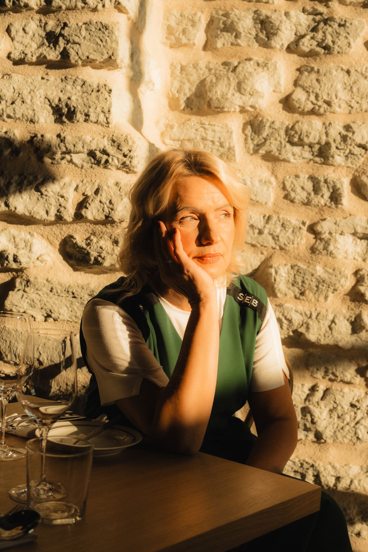 A person in a green vest sits thoughtfully at a table against a stone wall in warm lighting.