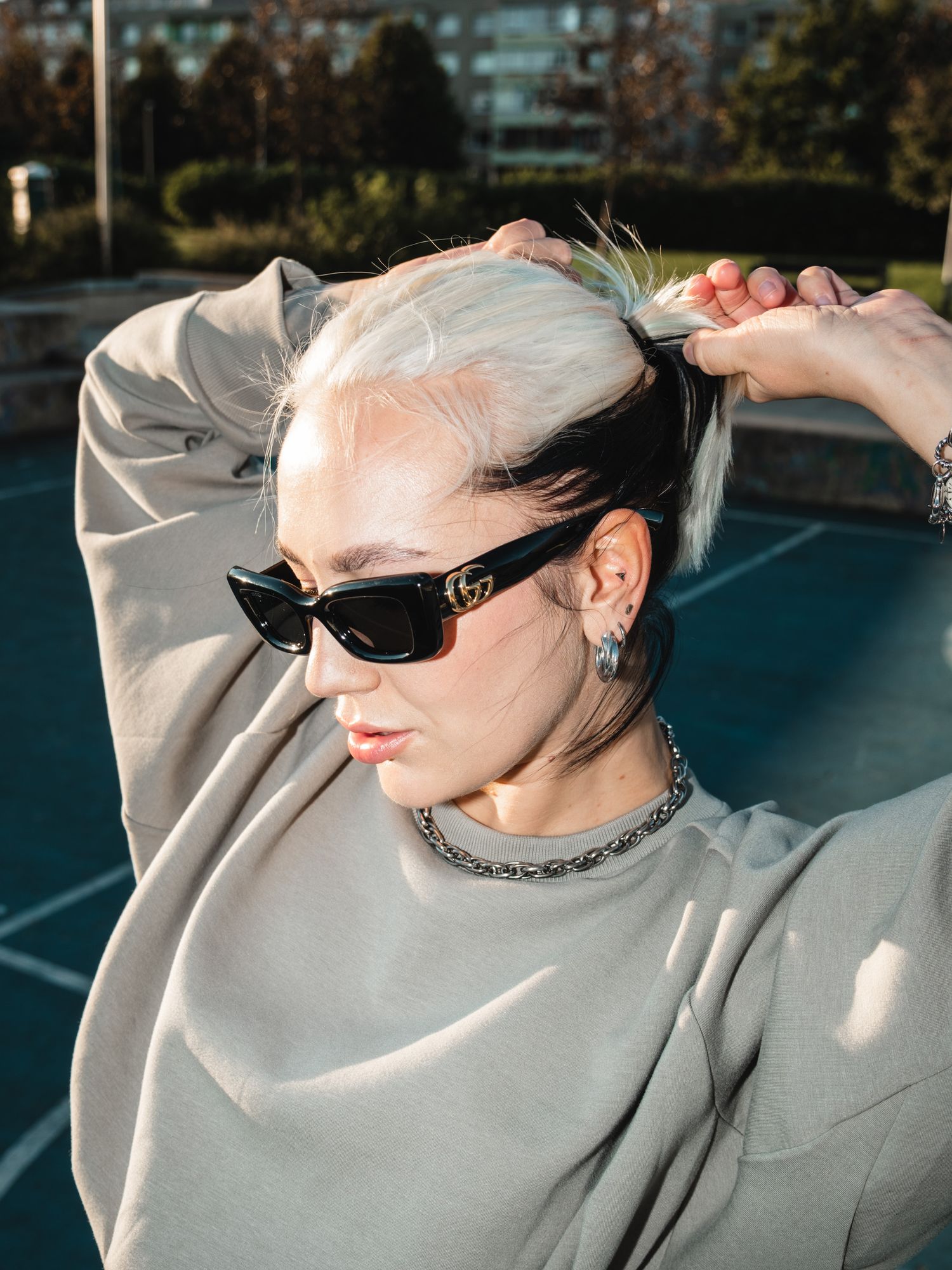 Person in grey sweatshirt and sunglasses posing in dramatic sunlight on outdoor court.