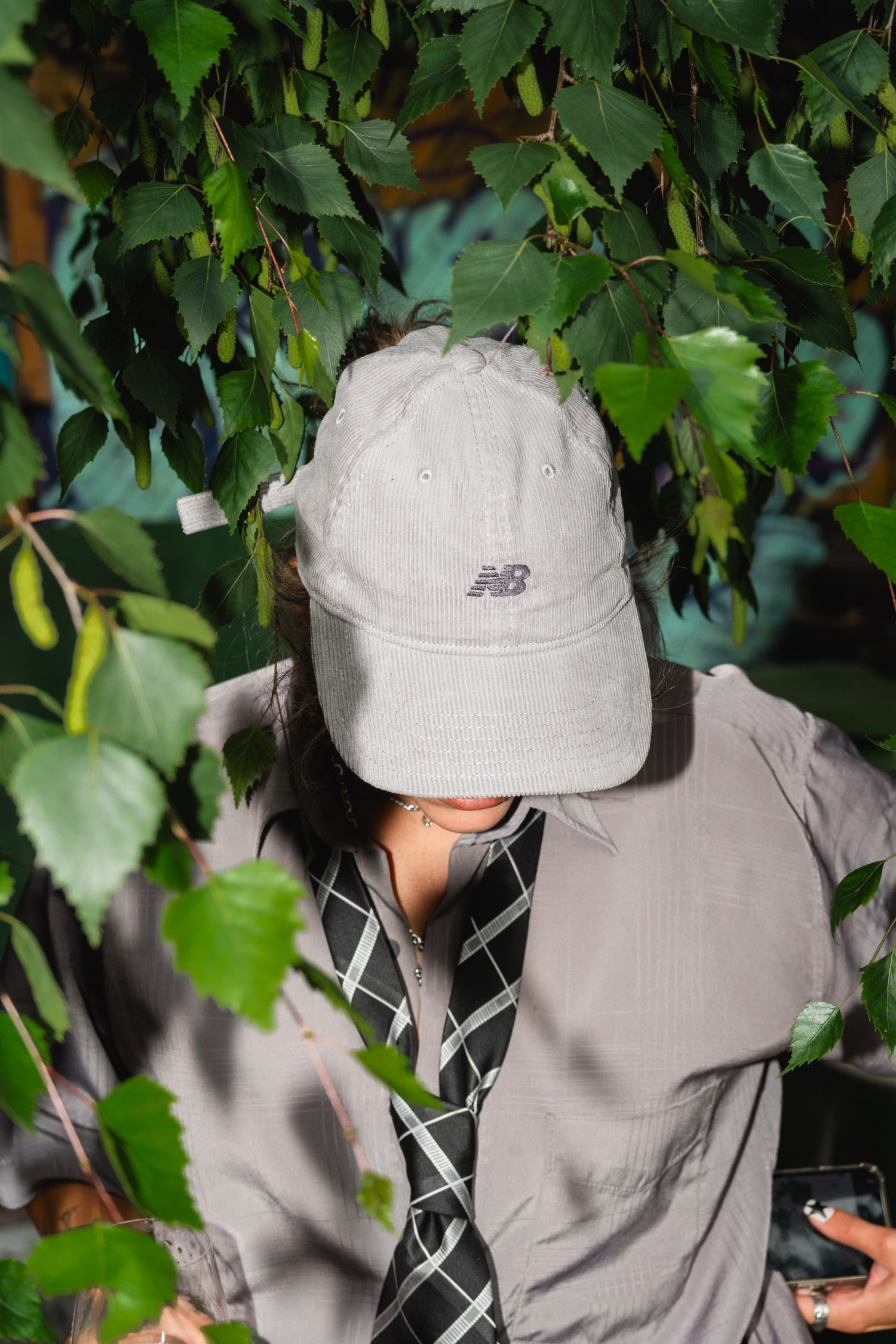 A person in a gray New Balance cap and jacket stands among green leaves.
