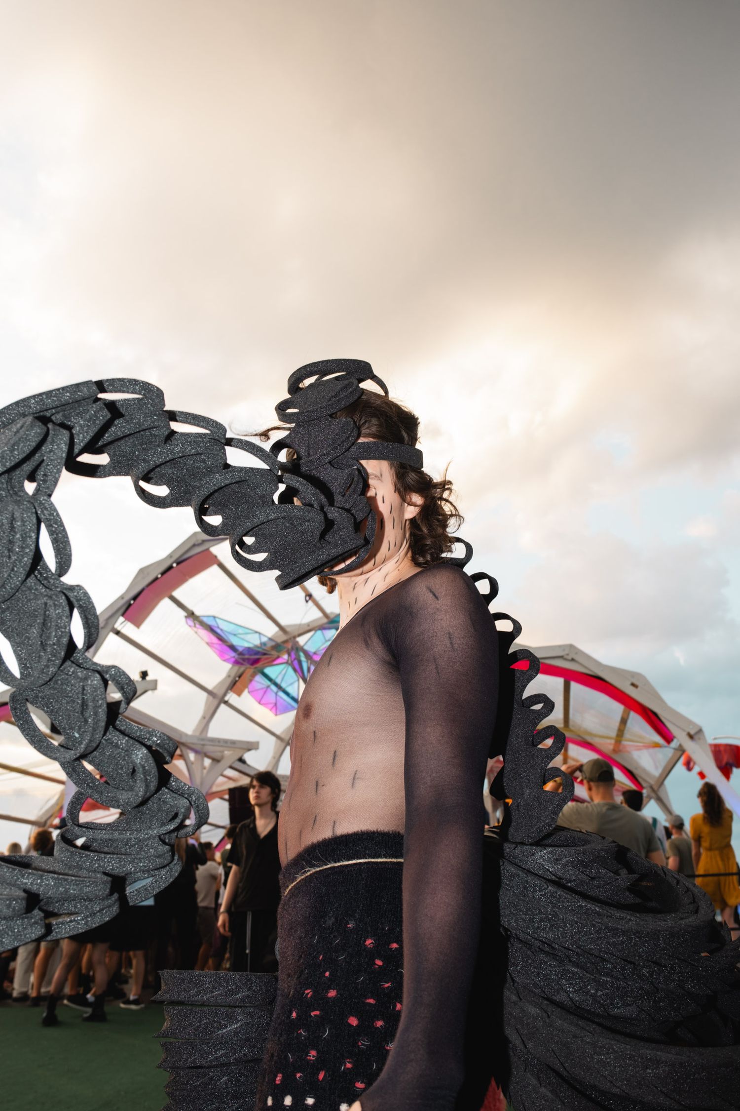 Festival attendees in dark costumes with decorative wings against cloudy sky.