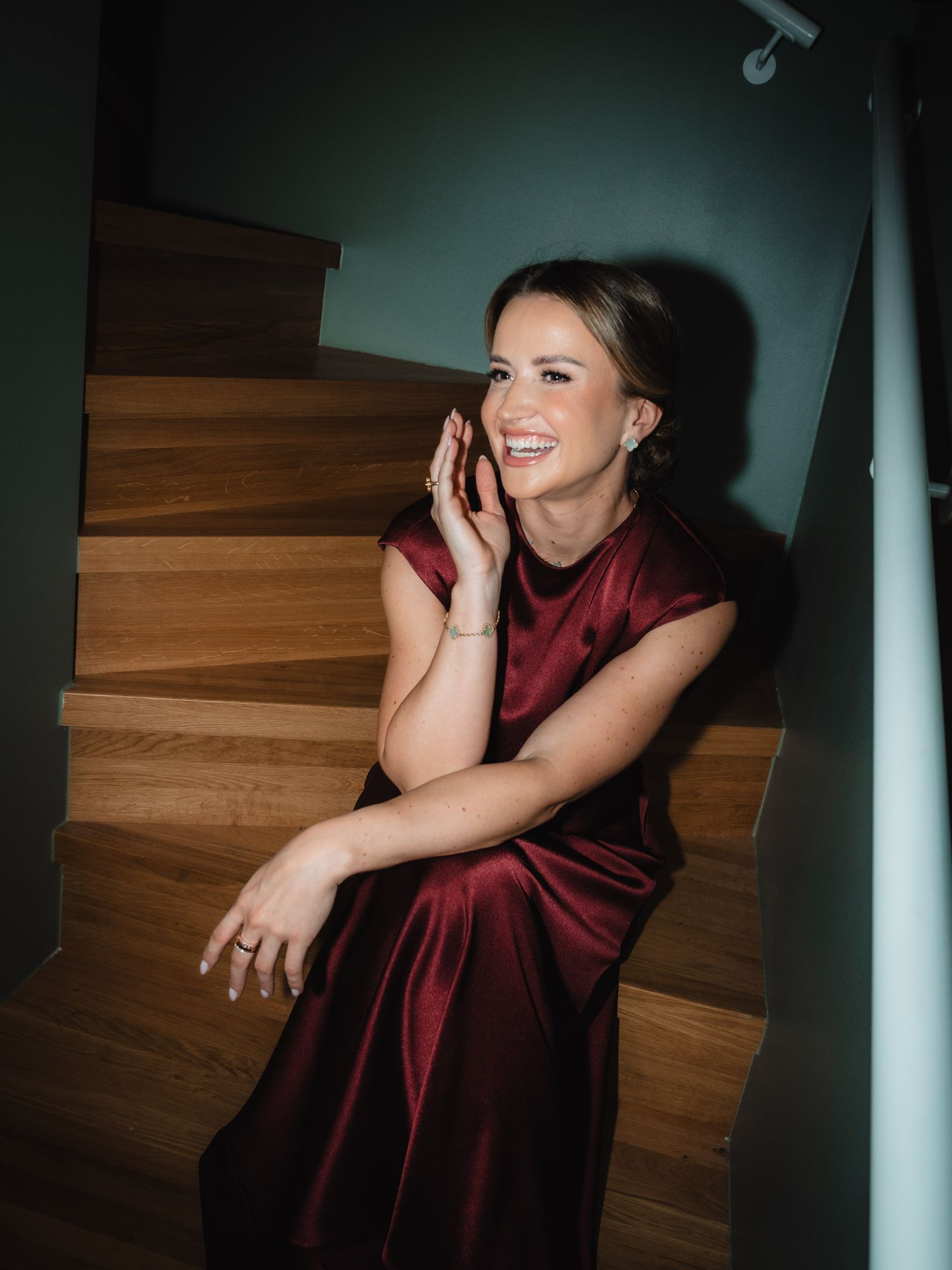 A person in a burgundy velvet dress sits on wooden stairs while smiling and talking on a phone.