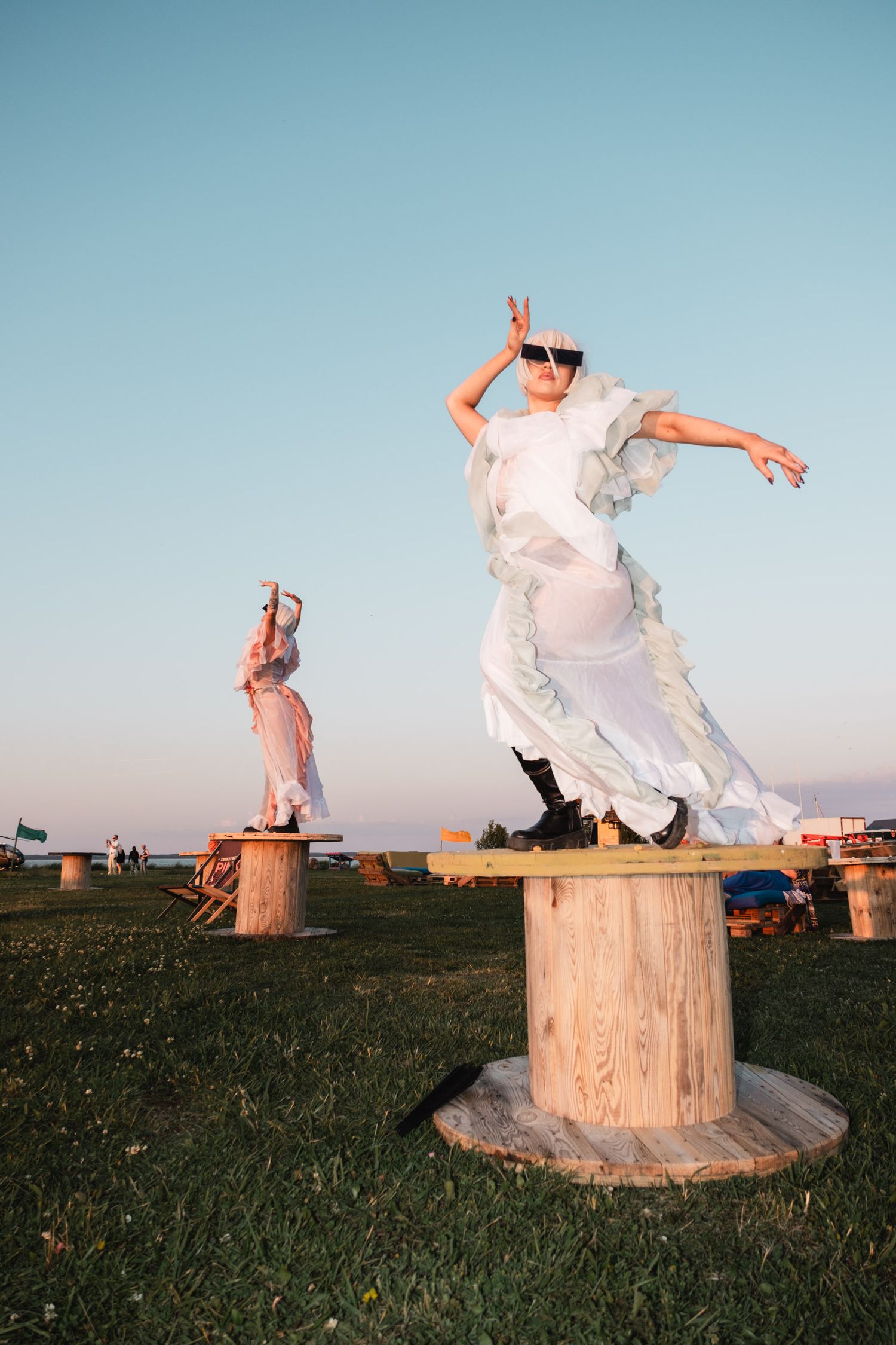 Person in flowing white dress performing artistic movements on wooden stumps at sunset.