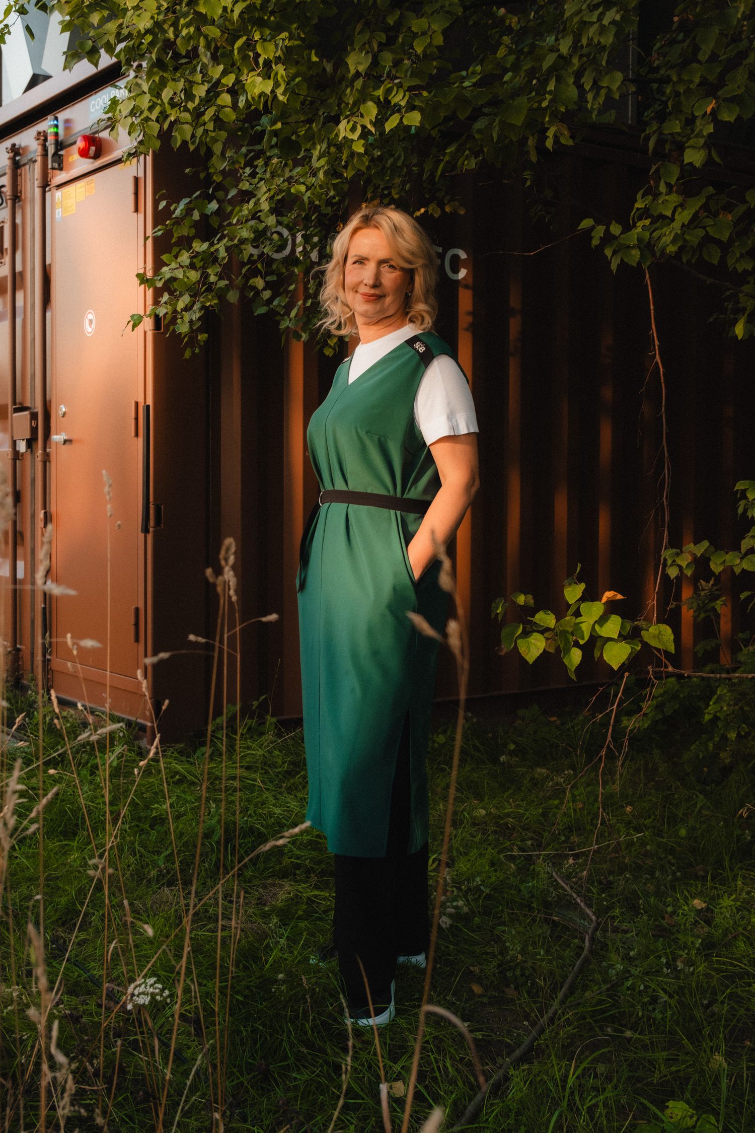 Person wearing a green apron stands in a lush garden at dusk with warm lighting in the background.