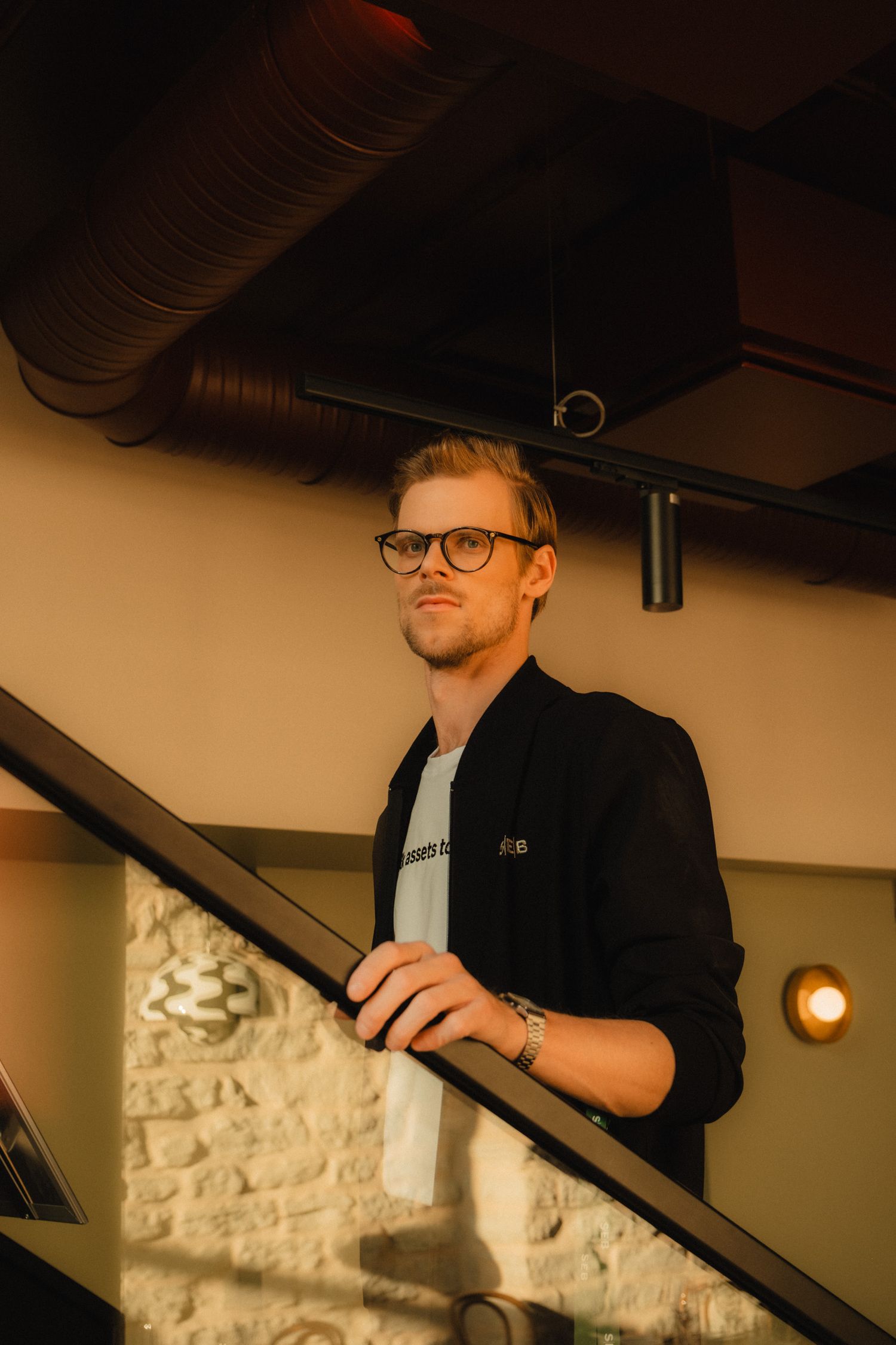 A person in glasses and dark clothing stands on a staircase with exposed stone walls in warm lighting.