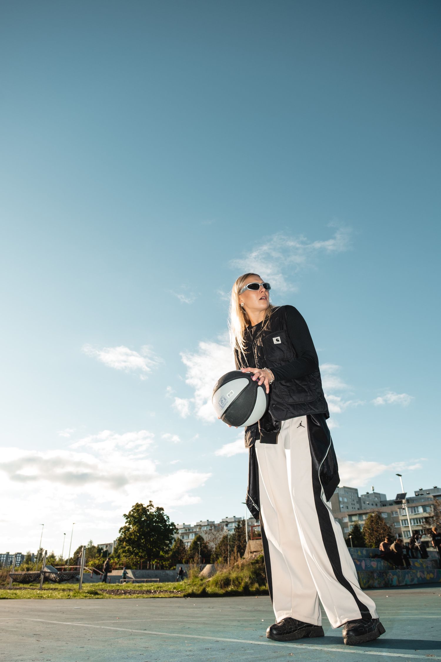 Person in black jacket and white pants holding ball against blue sky.