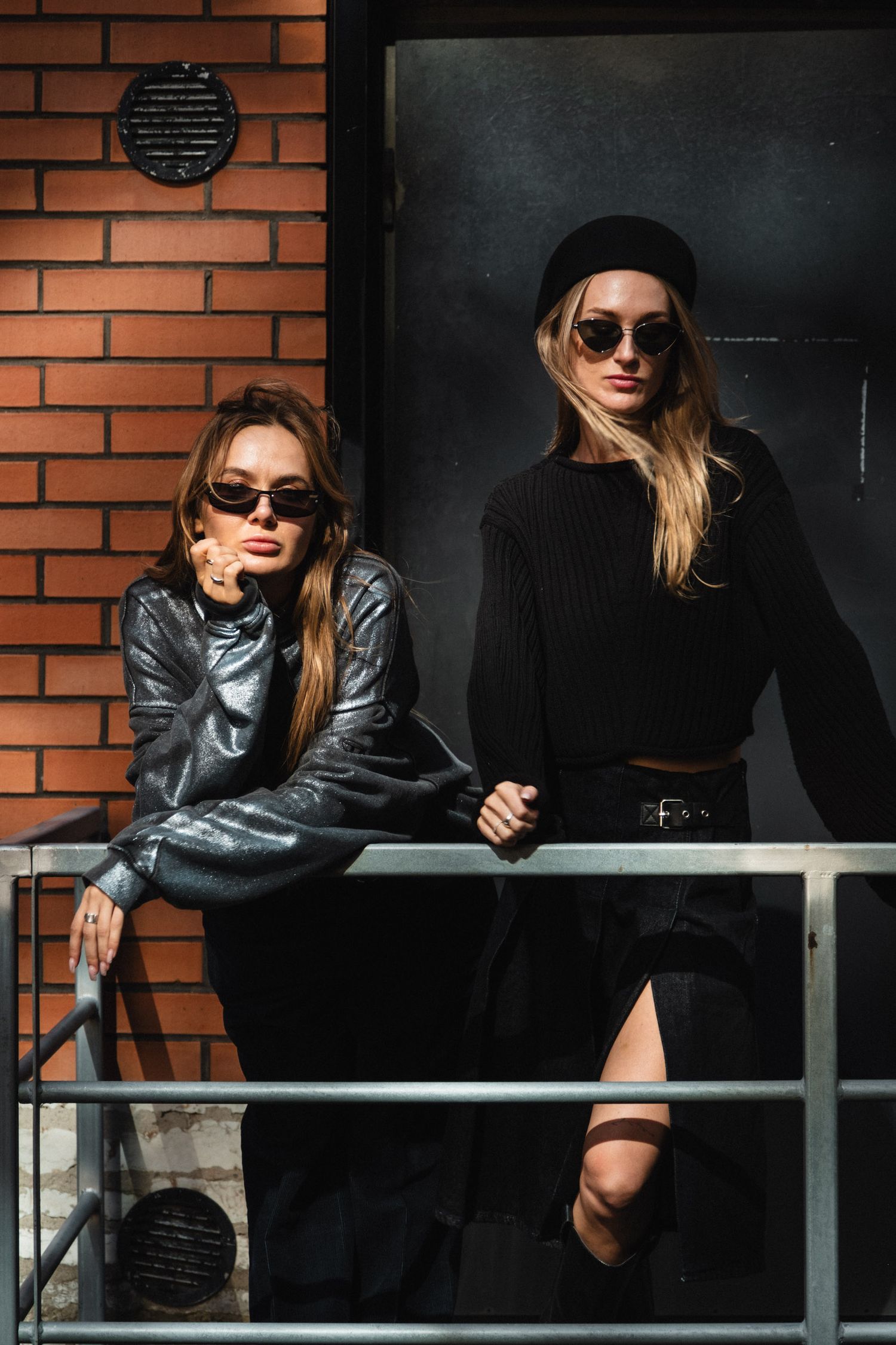 Two stylish women in black outfits and sunglasses posing by a metal railing against a brick wall.
