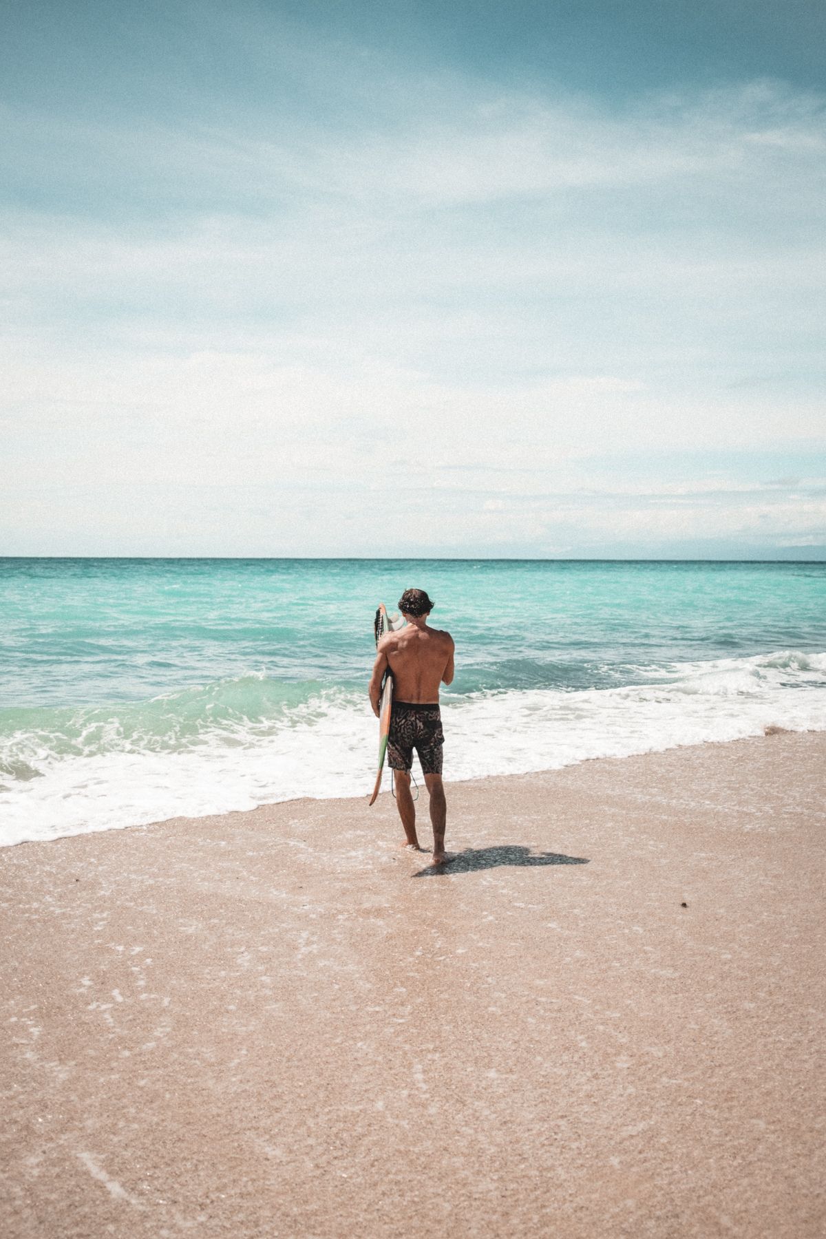 Solitary figure standing on sandy beach looking out at turquoise ocean waves.