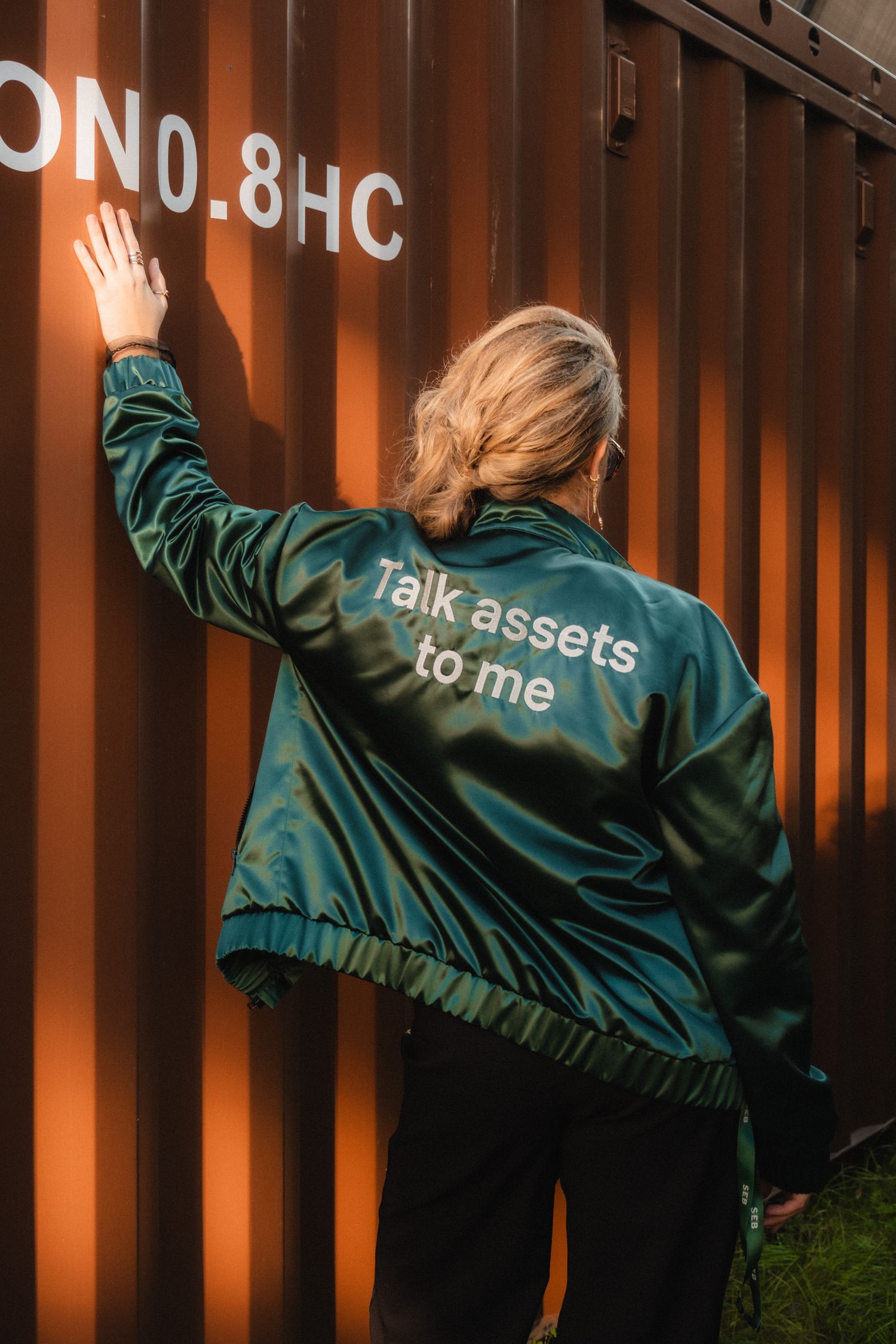 Person wearing green satin jacket with 'Talk assets to me' text stands against orange shipping container in outdoor setting.
