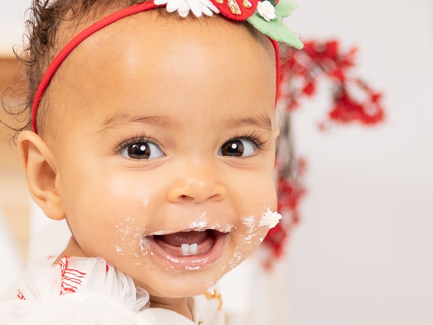 Close-up portrait of a baby wearing a red and white floral headband against a white background.