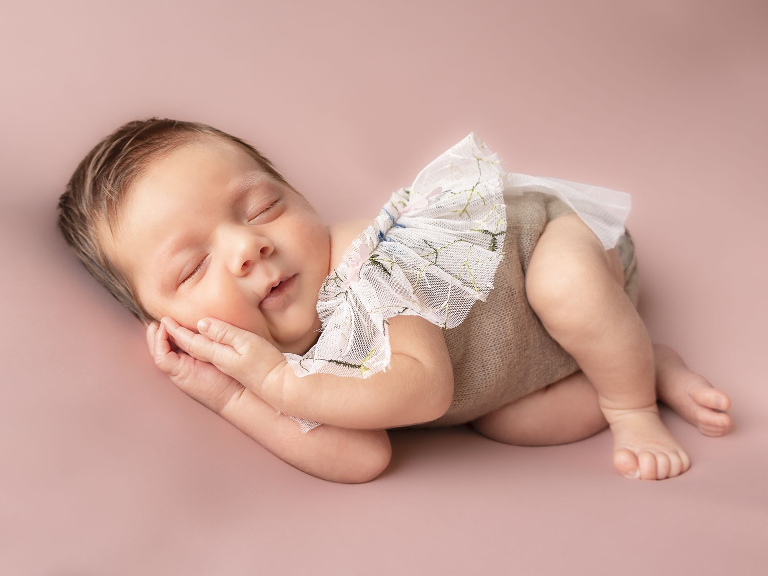 Sleeping newborn in white wrap photographed against soft pink backdrop.