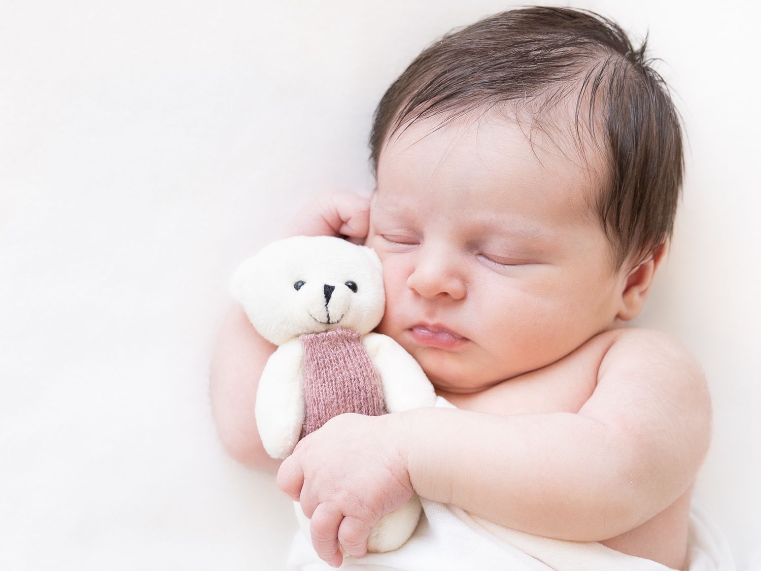 Sleeping newborn cuddling small teddy bear on white background.