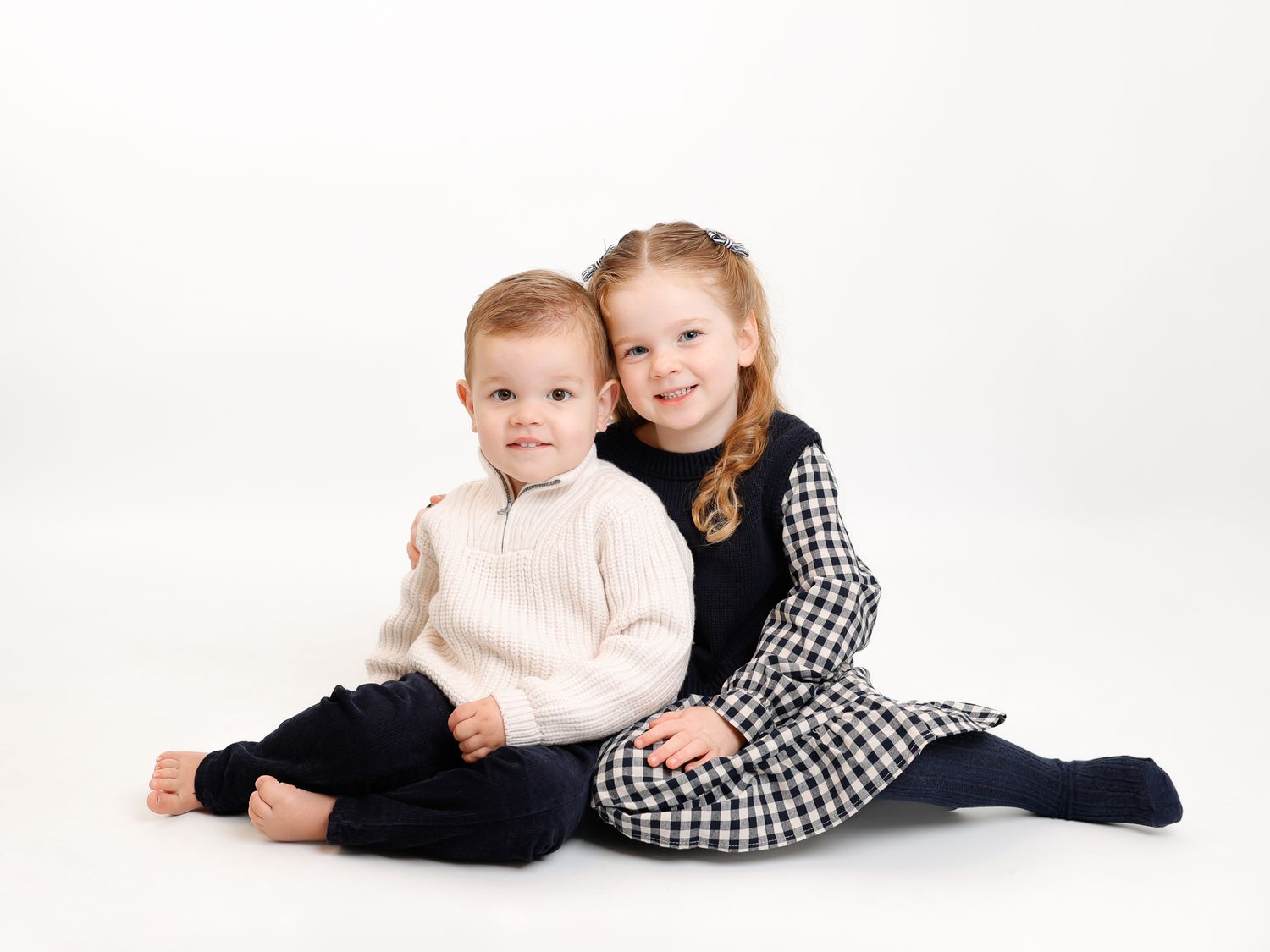 Two young siblings pose together smiling, boy in cream sweater and dark pants, girl in checkered dress, on white background.