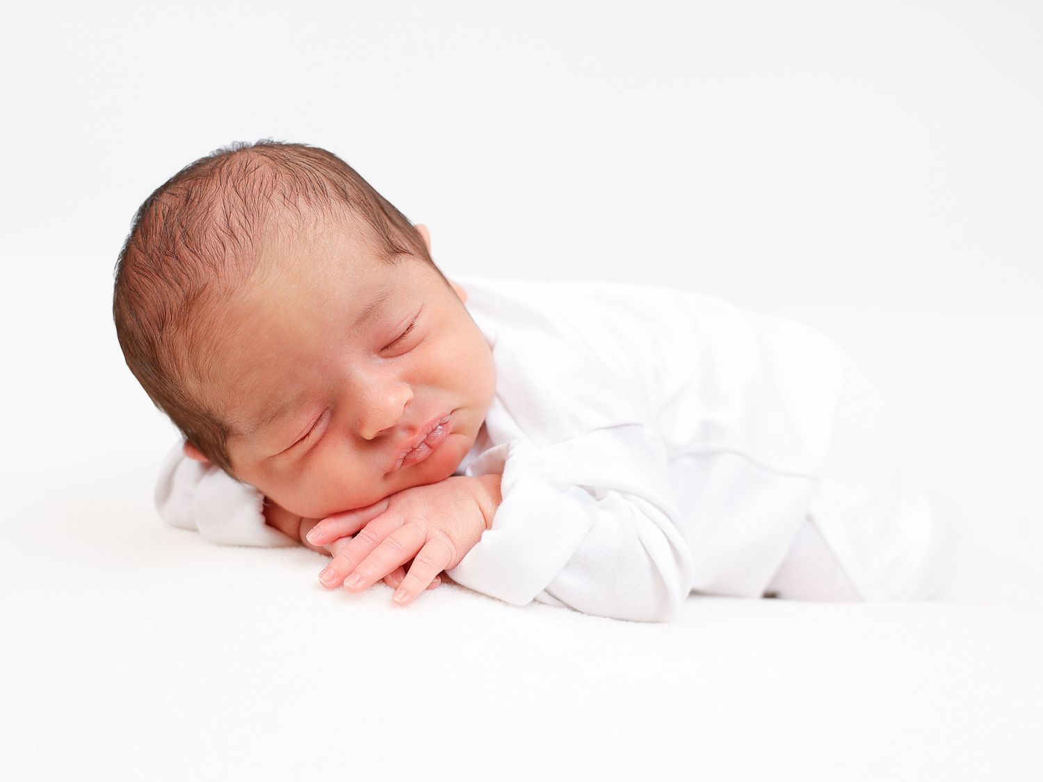 Peaceful newborn baby sleeping in white outfit on white background, curled up with hands under chin.