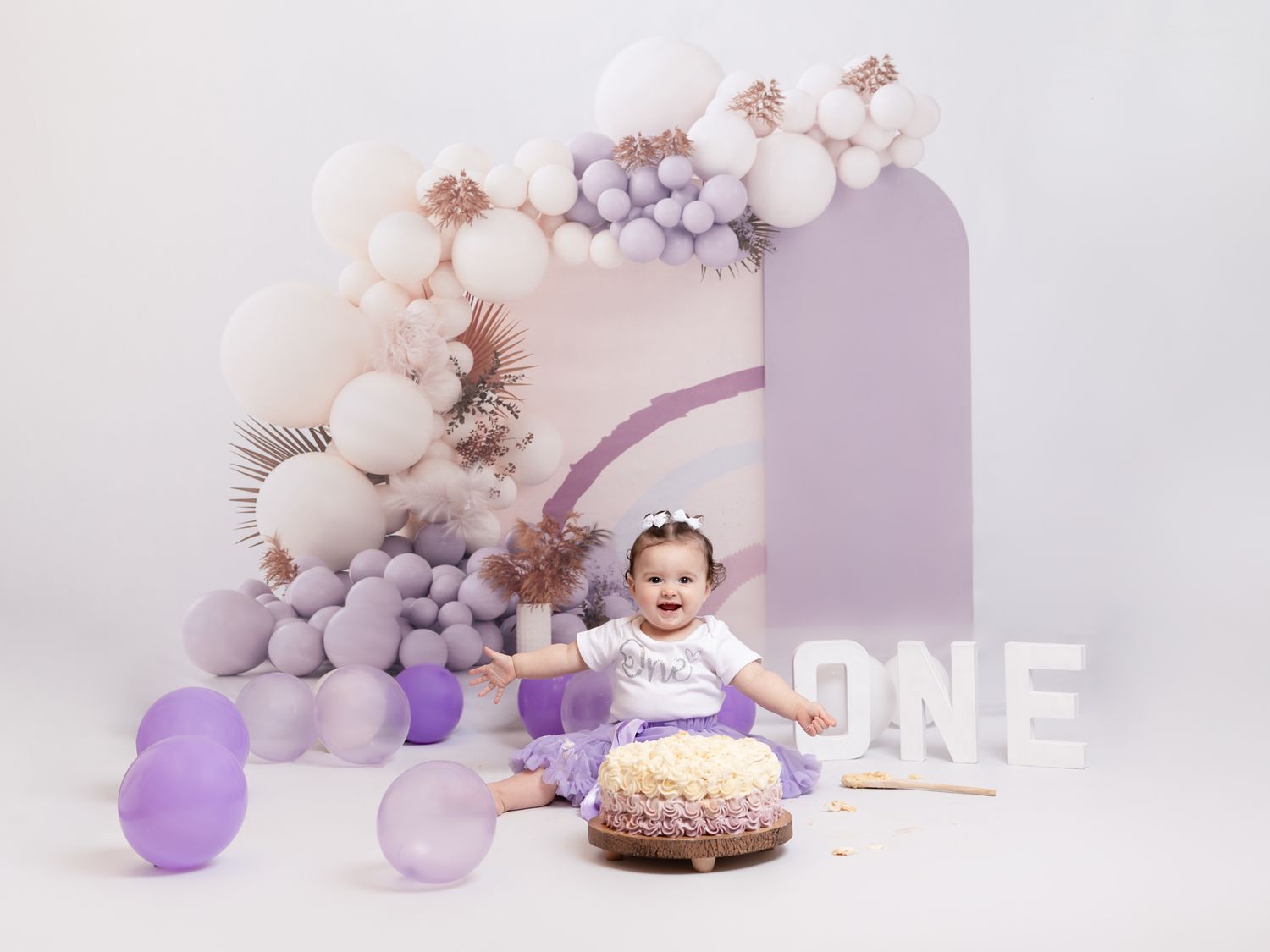 First birthday cake smash session with purple balloon arch and white backdrop.