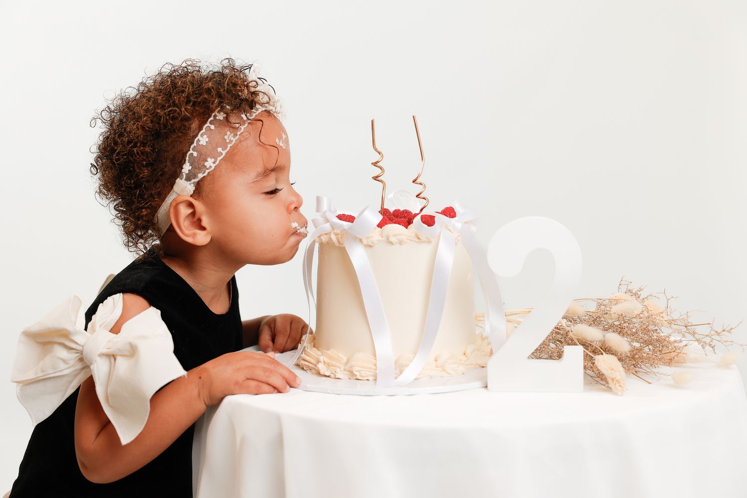 Toddler girl in black dress blowing out candles on a white birthday cake celebrating her 2nd birthday.