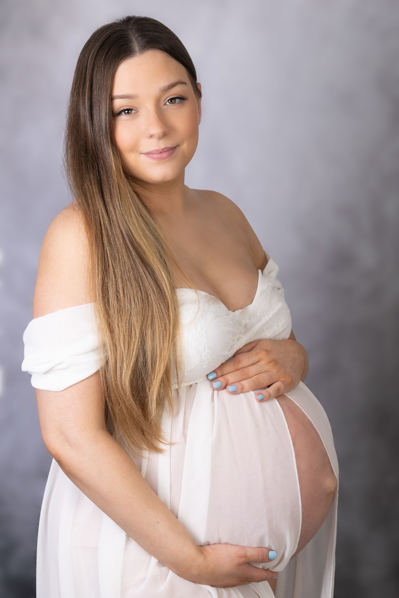 Maternity portraits of woman in white off-shoulder dress against grey background.