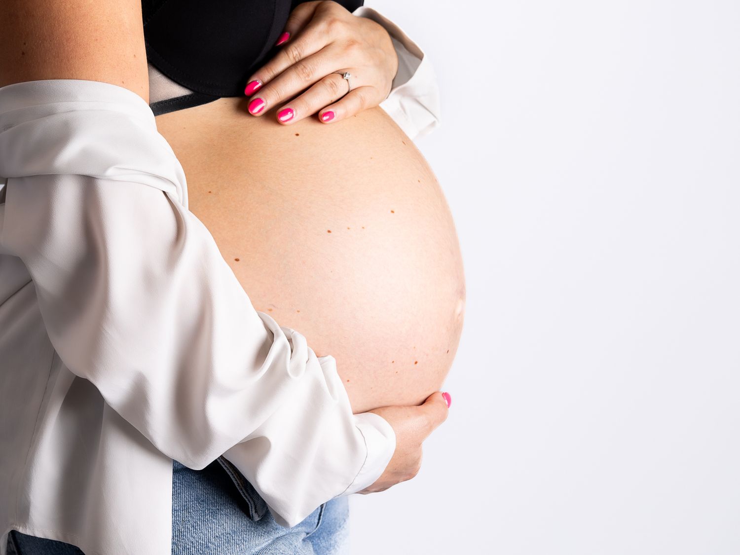 Expectant mother in a white shirt caressing her pregnant belly against a light backdrop.