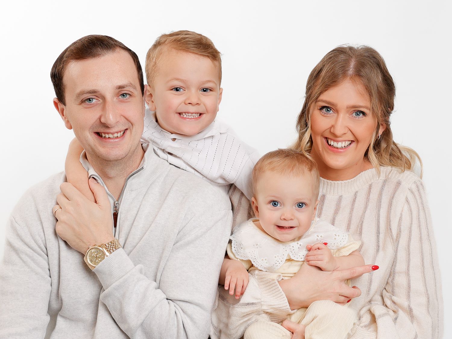 Happy family portrait with parents holding two young children, smiling against a white background.