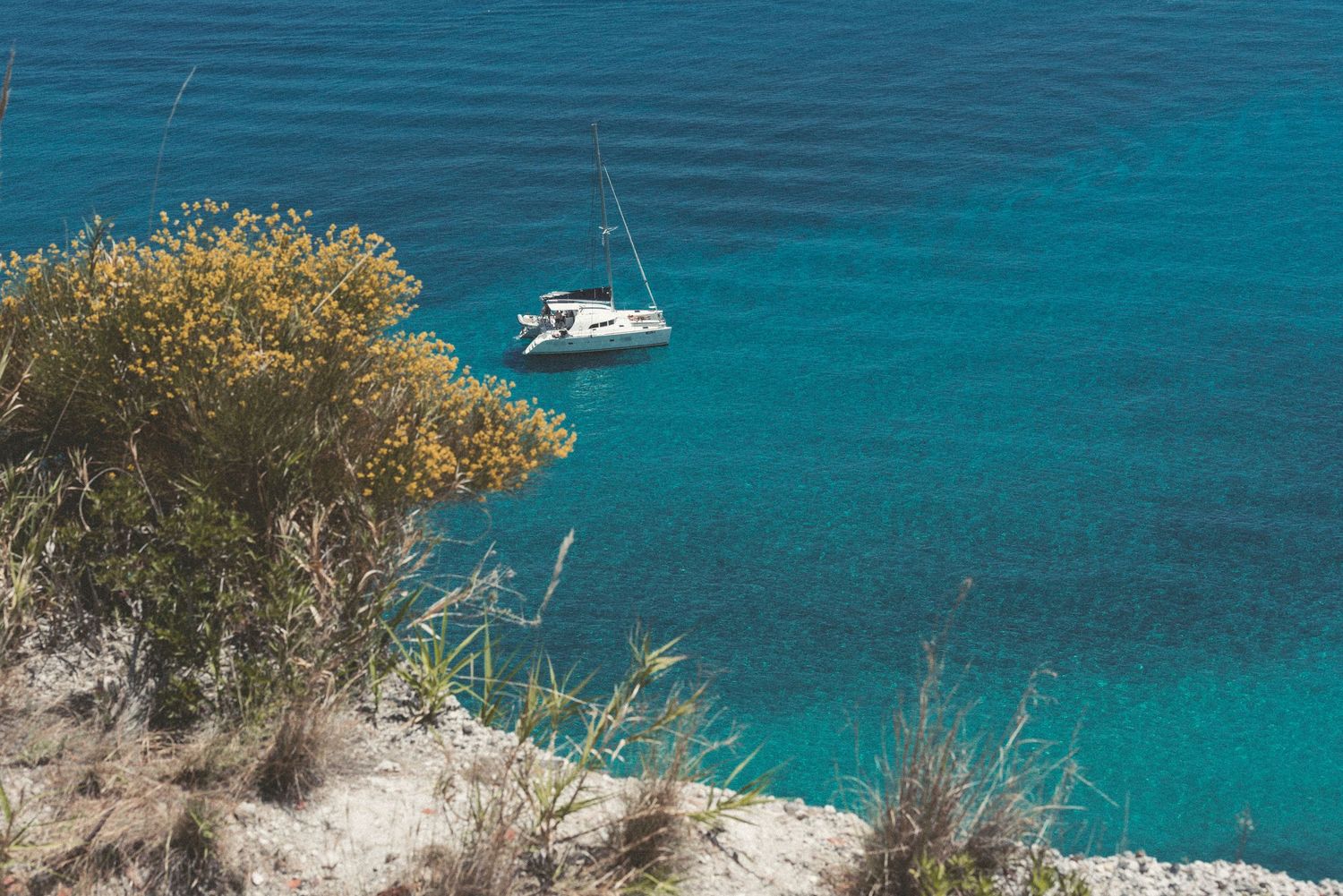 Aerial view of turquoise Mediterranean waters with boats anchored near rocky coastline and green vegetation.