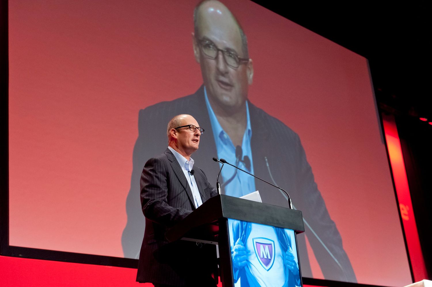 Technology conference presentation with TV personality David Koach showing on multiple screens against red backdrop.