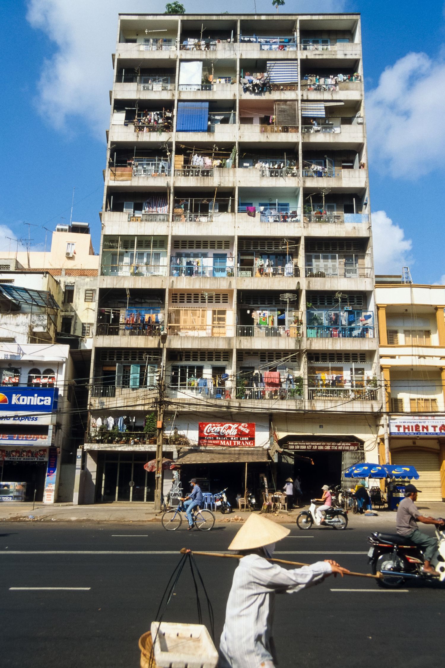 Multi-story apartment building with balconies and storefronts along a street in an urban setting.