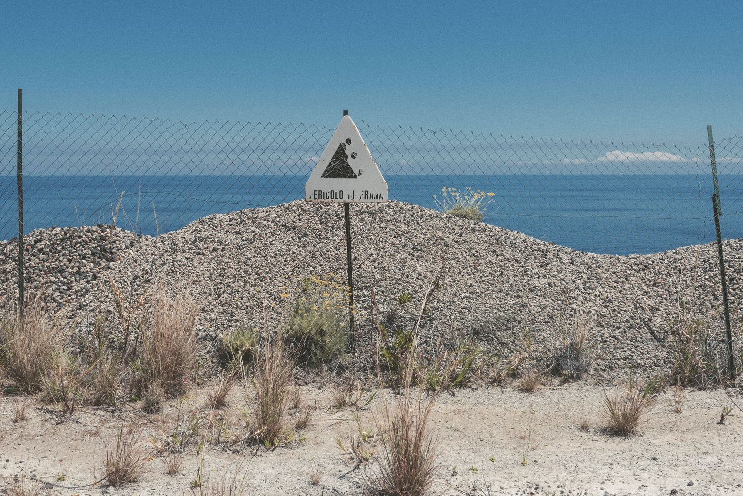 Warning sign stands against blue sky on sandy beach with chain link fence and desert vegetation in foreground.