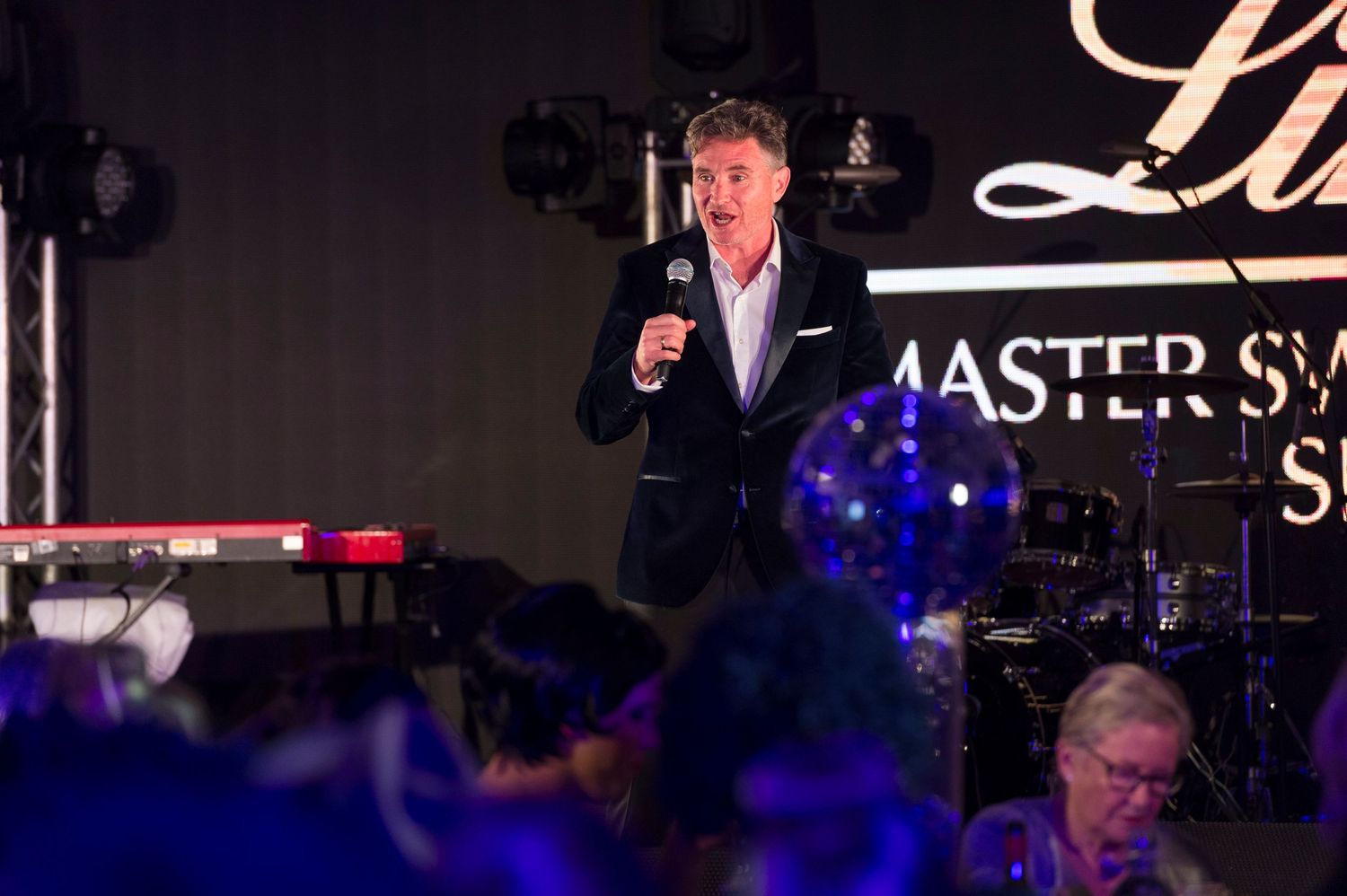 A speaker in a dark suit addresses an audience at a Master Sync event with blue lighting and stage decor.