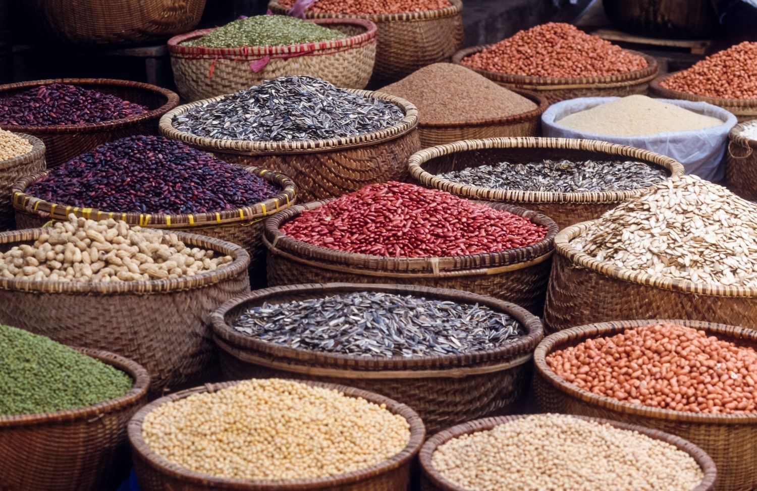 Colorful assortment of dried beans and grains displayed in round woven baskets at a market.