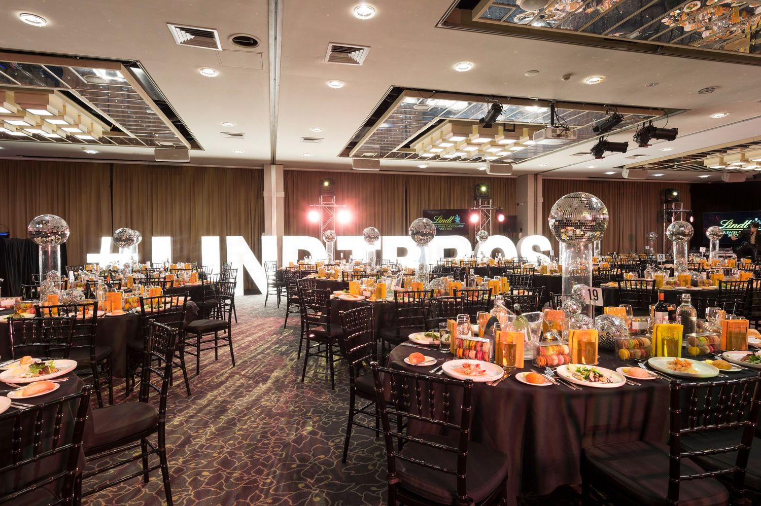 Large banquet hall set up for an elegant event with round tables, brown linens, and orange floral centerpieces.
