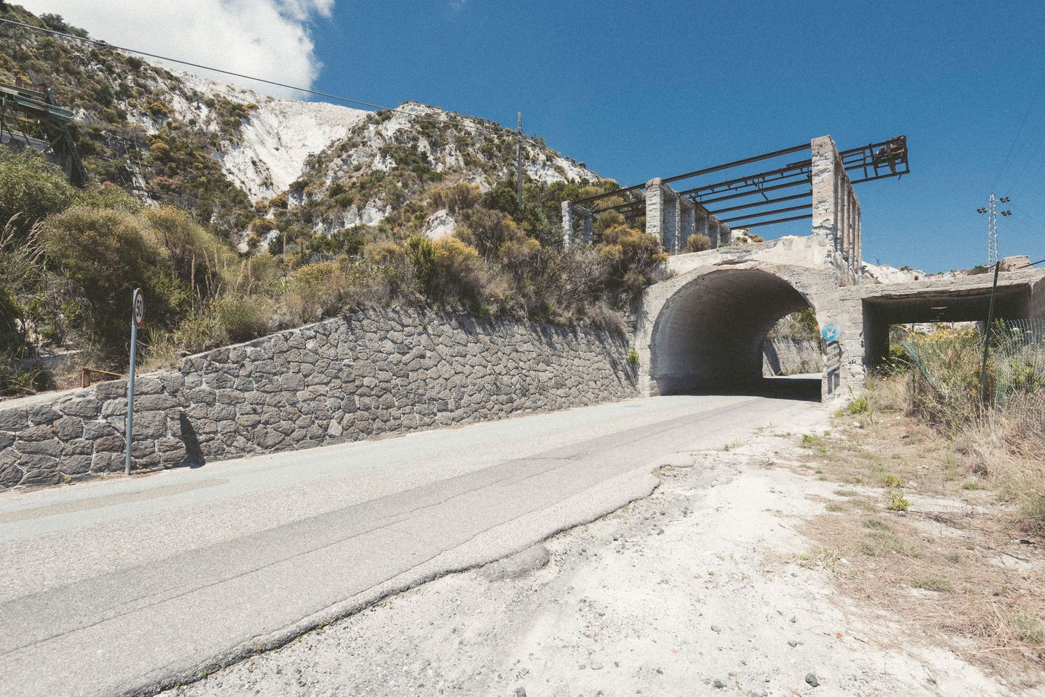 Winding concrete road leading up a steep hillside with stone retaining walls and coastal views.