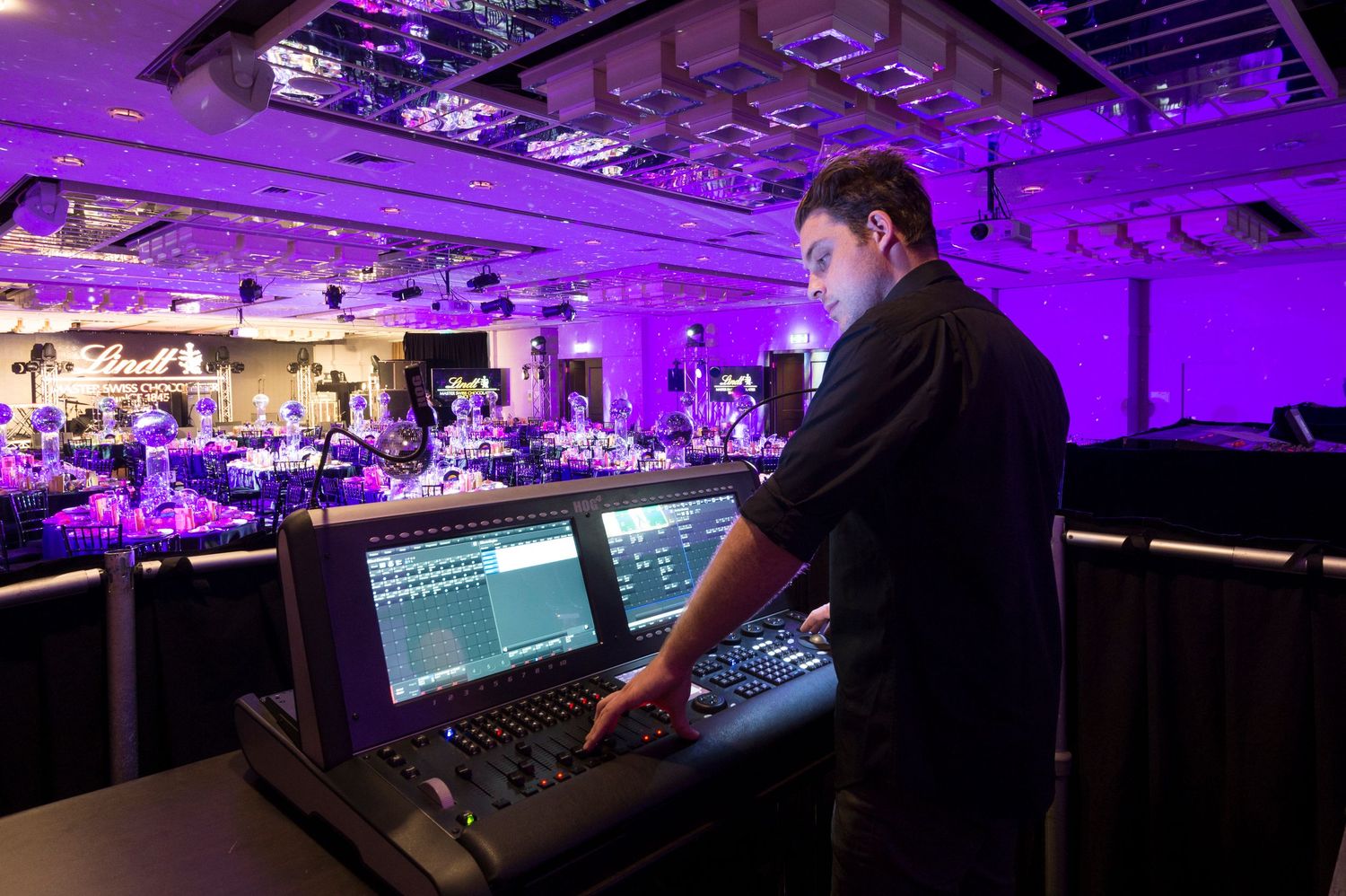 Audio engineer operates a digital mixing console at an event venue illuminated with purple lighting.