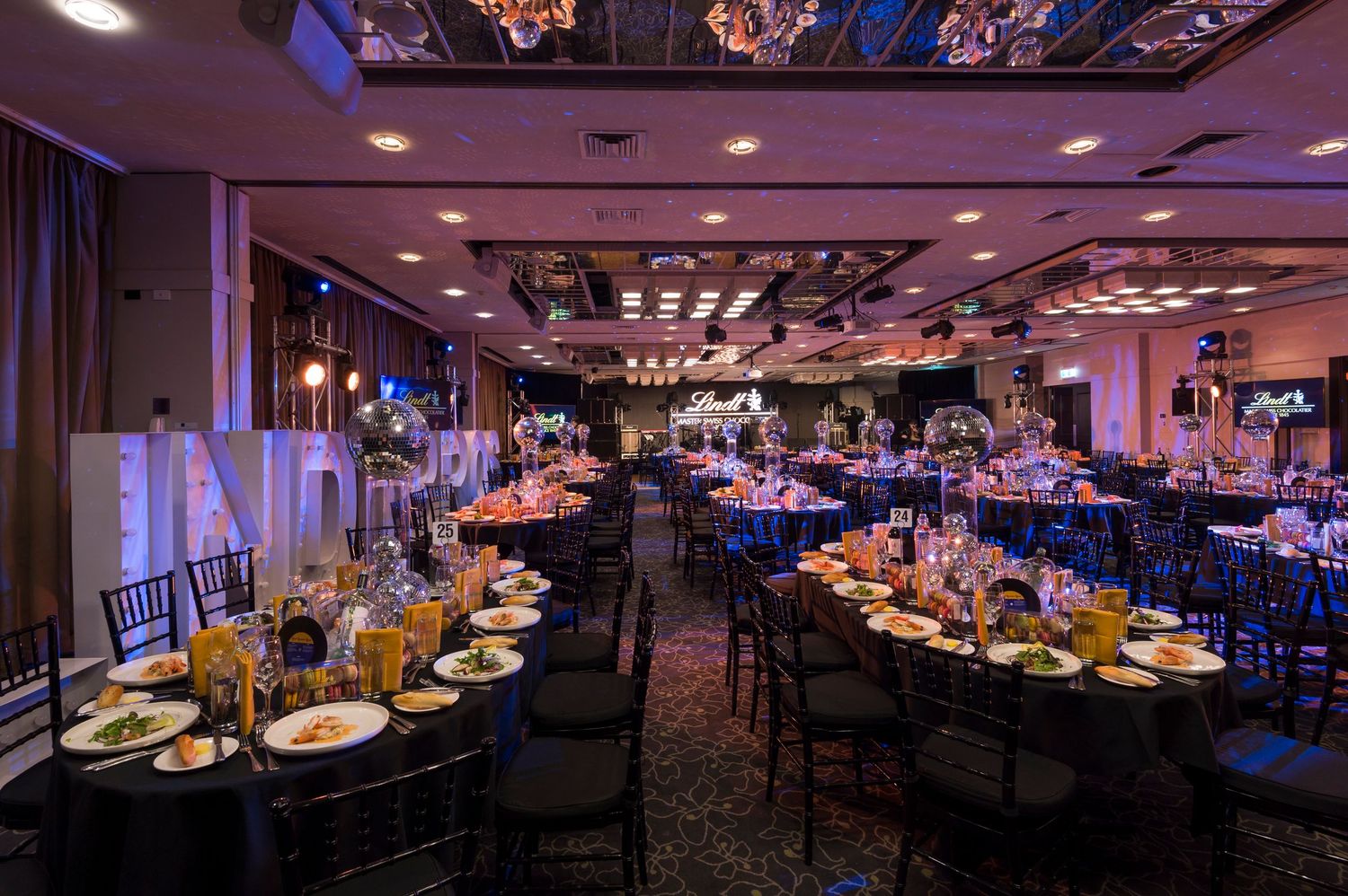 A dimly lit banquet hall with elegant table settings, purple ambient lighting, and multiple dining areas extending into the distance.