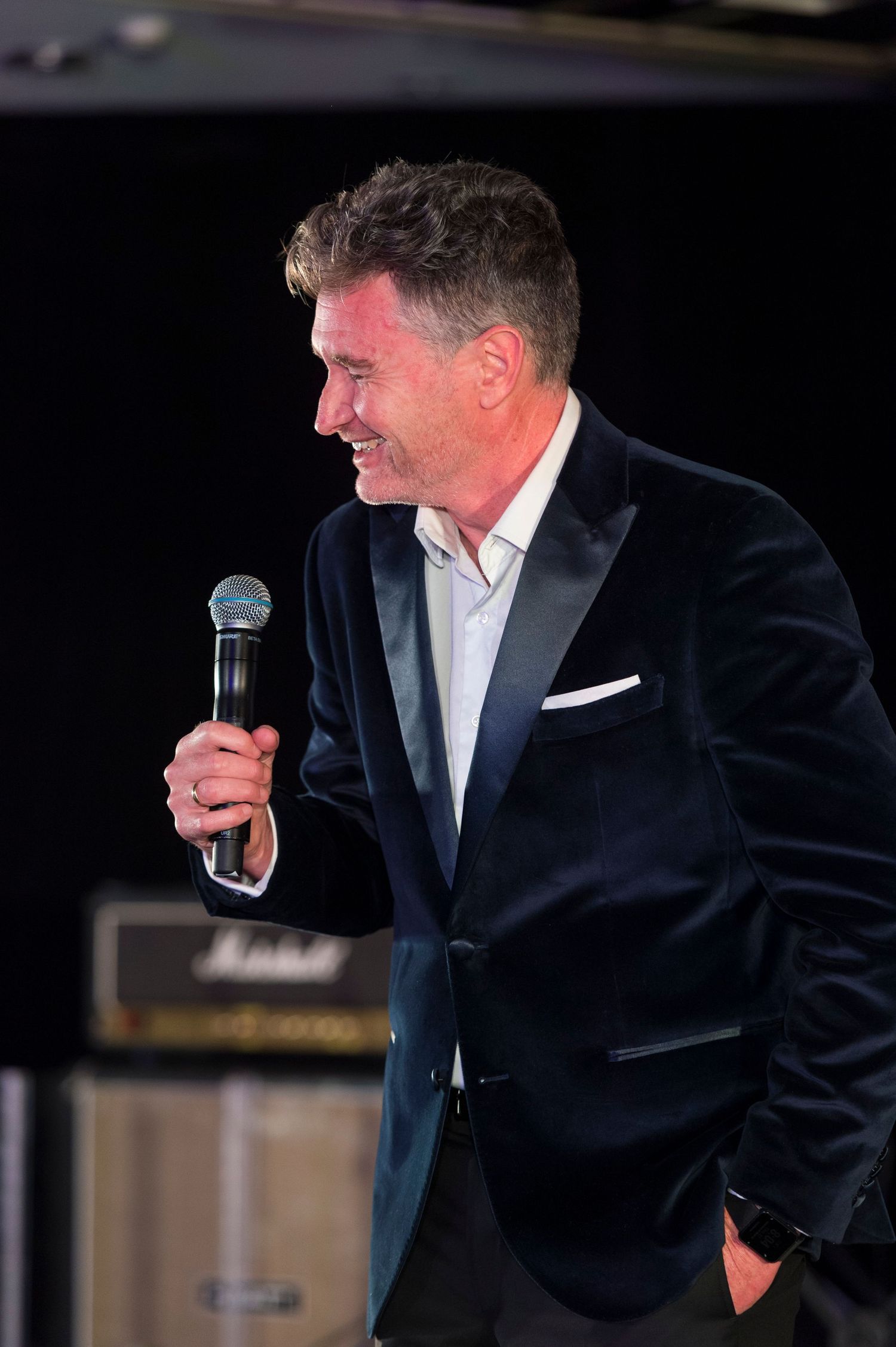 A performer in a black suit and white shirt holds a microphone while gesturing expressively on stage during a show.