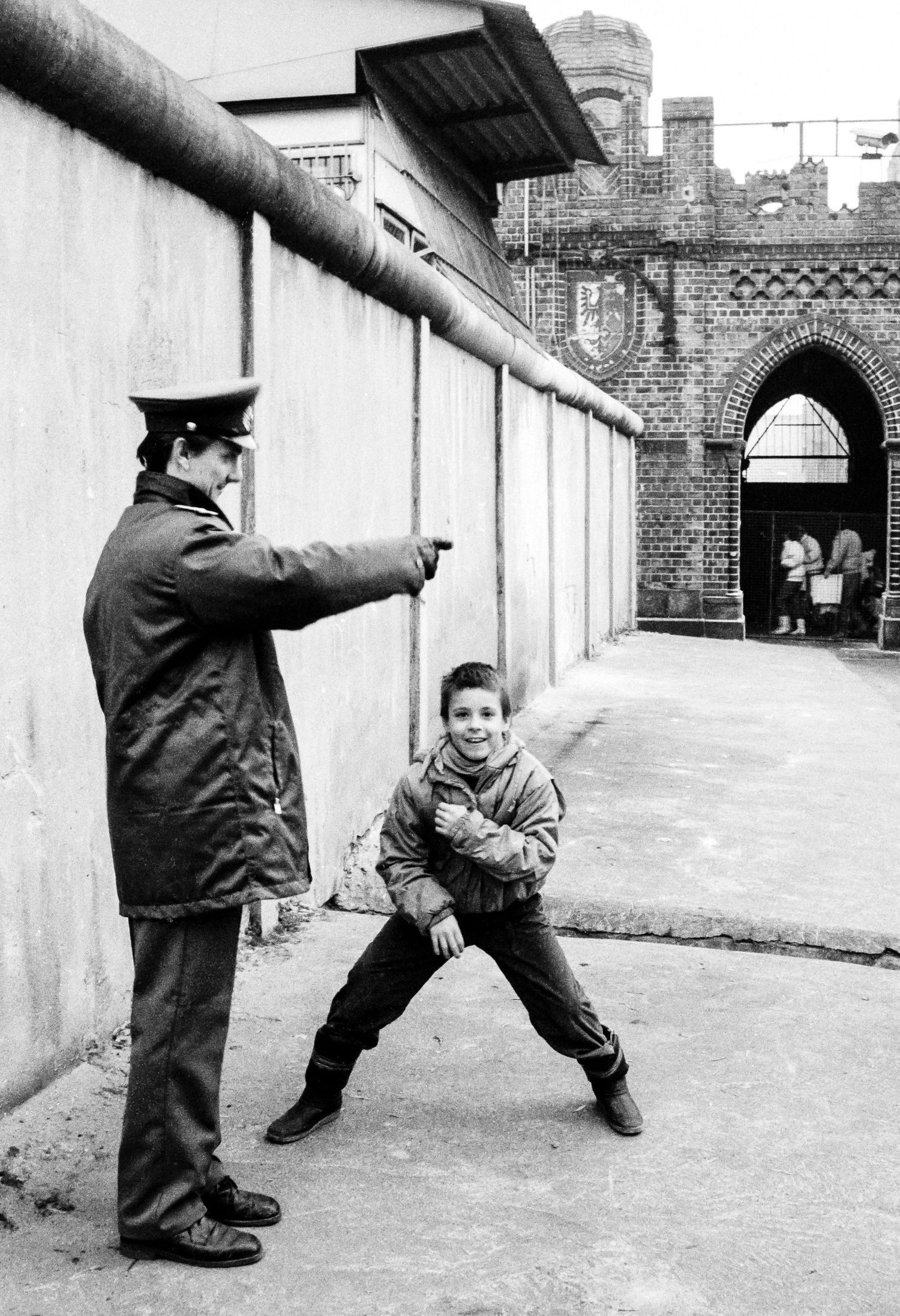 Children playing near the Berlin Wall with guard watching in black and white photo.