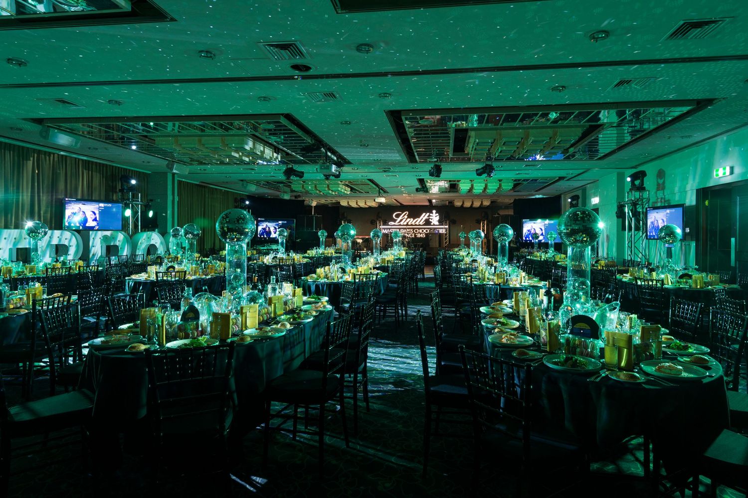 A styled banquet room set up with green lighting illuminates rows of tables and chairs set up for a formal event.