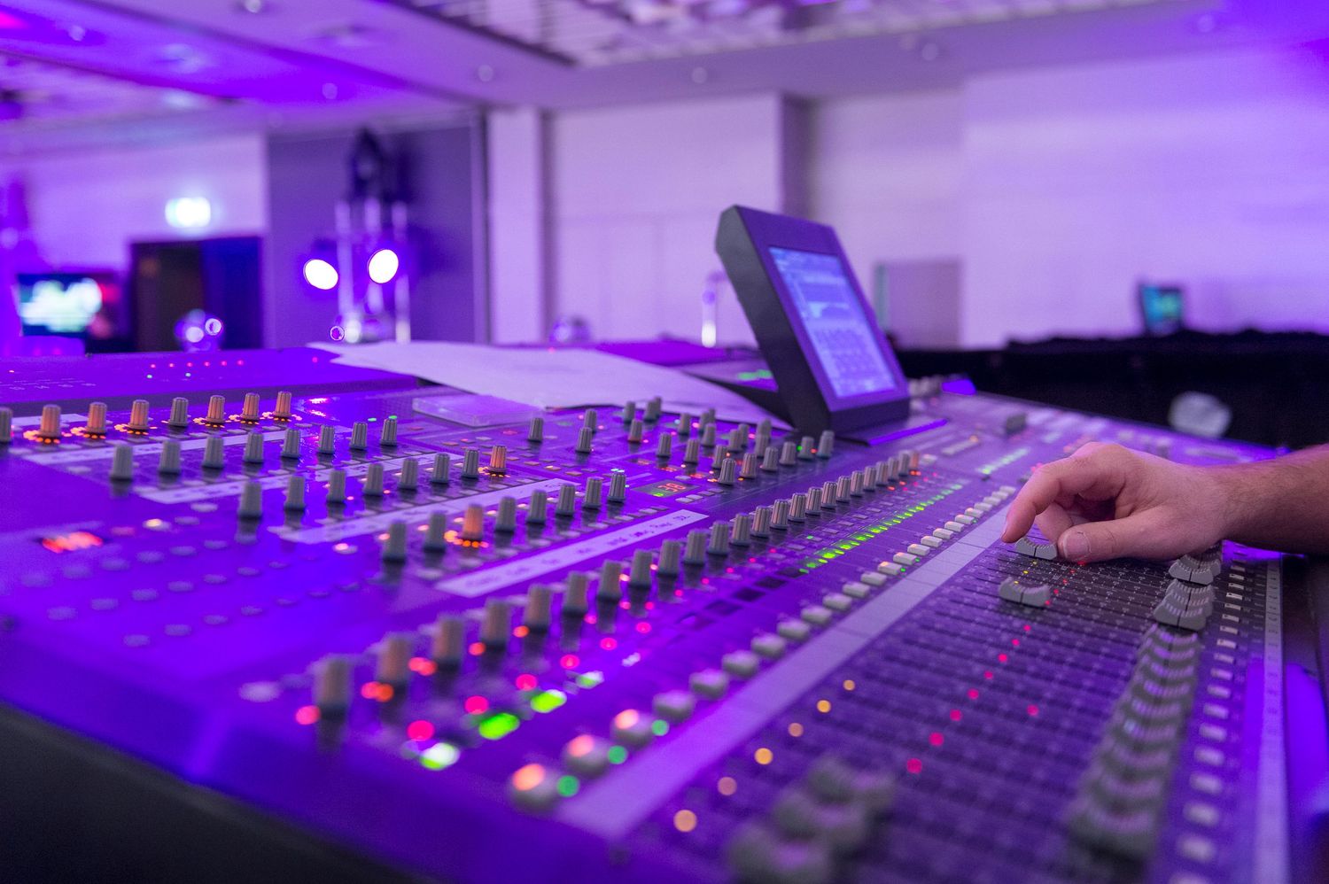 Professional staging audio-visual mixing console illuminated by purple stage lighting with hands adjusting controls in a music venue.