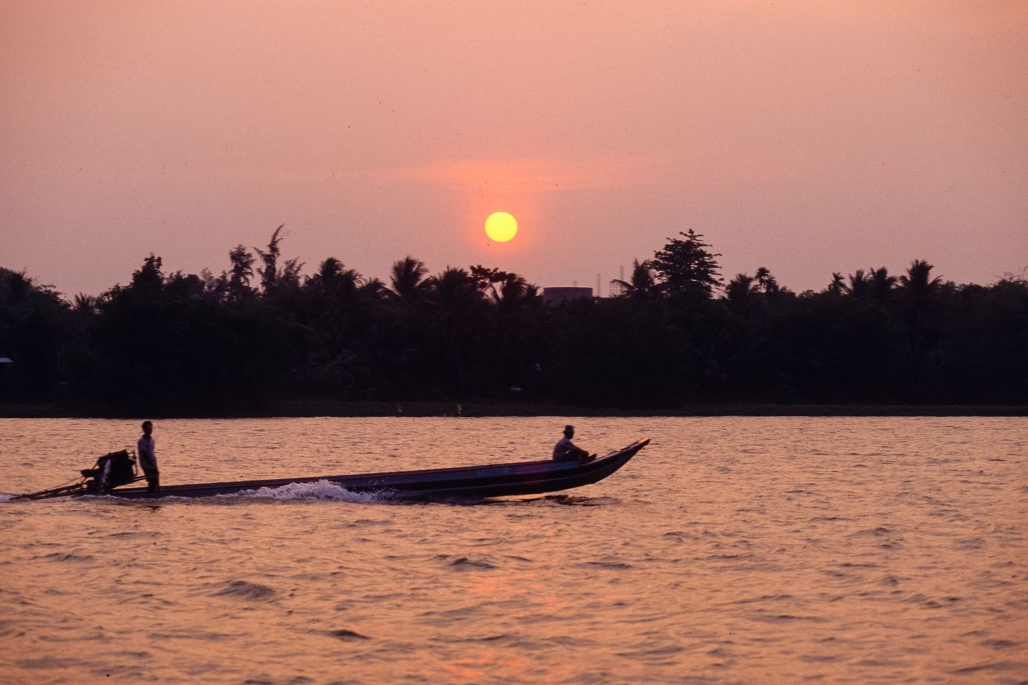 Boats silhouetted against a vibrant orange sunset over a tranquil river in Southeast Asia.