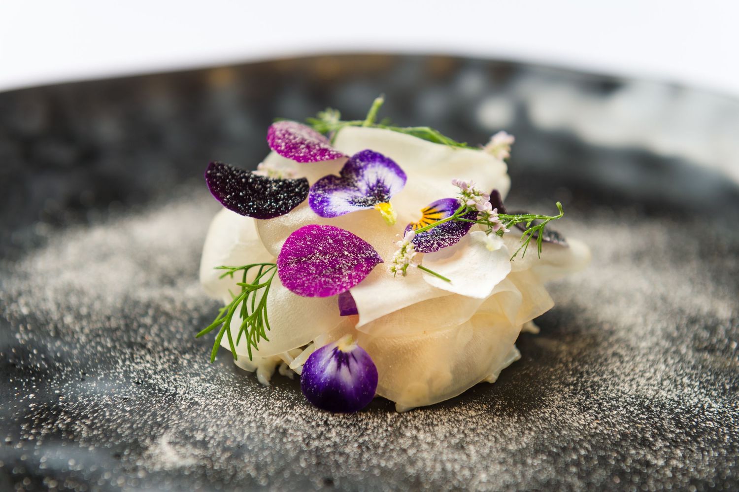 Elegantly plated dessert garnished with purple edible flowers and herbs on a dark textured surface.