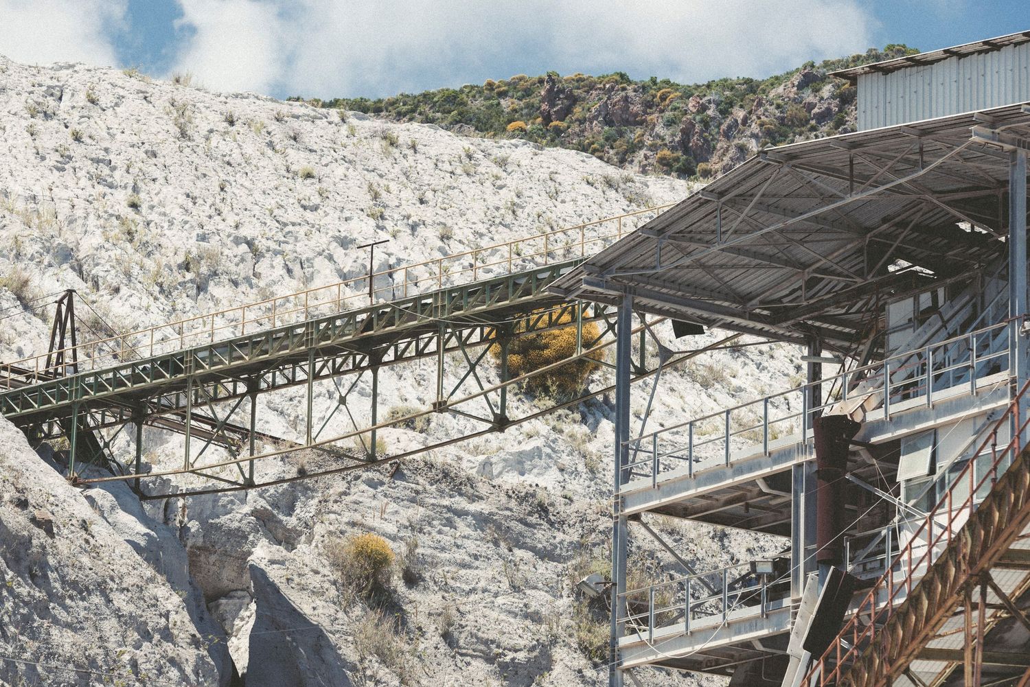 Industrial conveyor belt system and machinery operating in a white limestone quarry with rocky cliffs in the background.