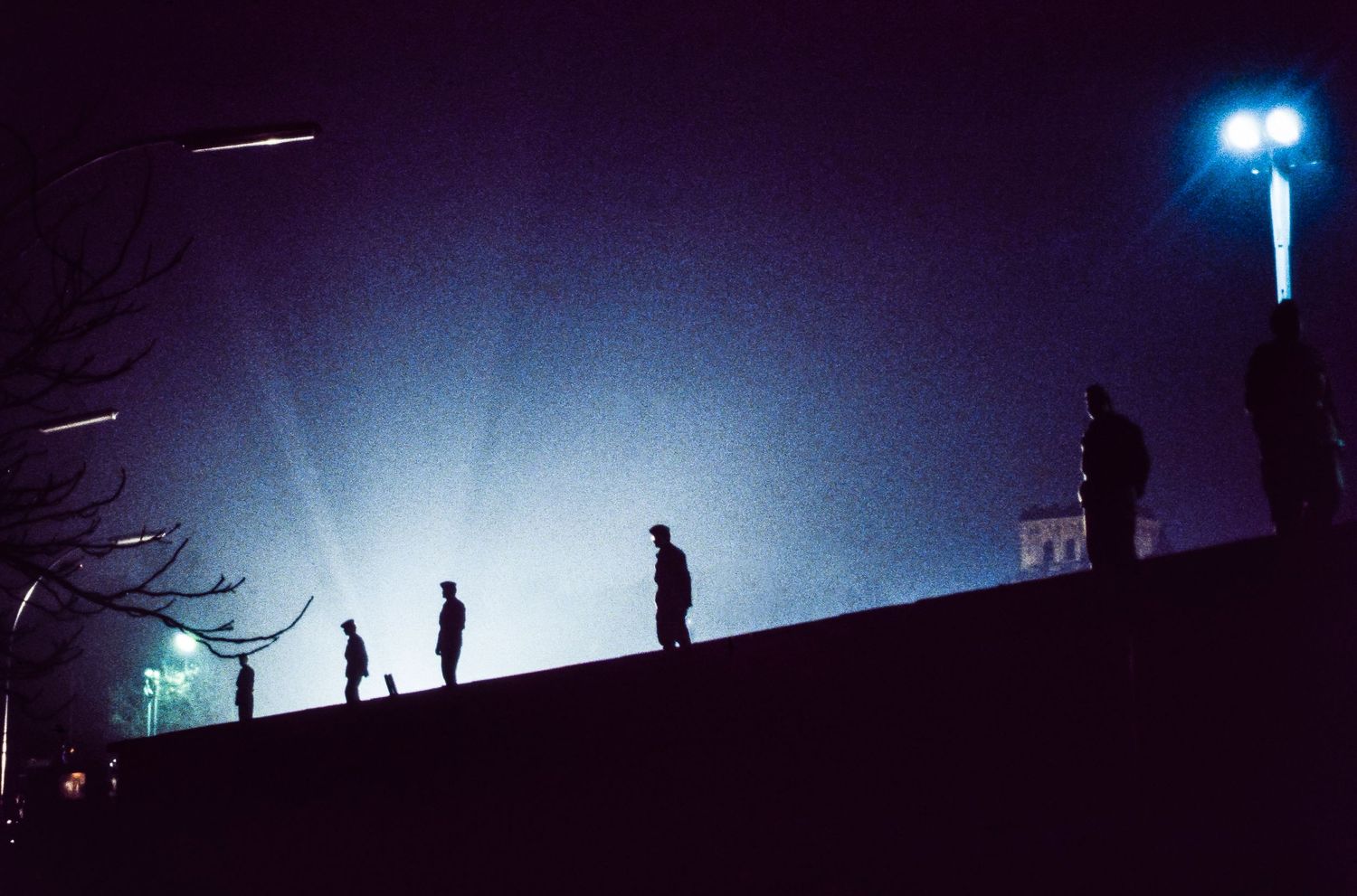 Dark silhouettes of people standing on a hill against a blue twilight sky with bright lights in the distance.