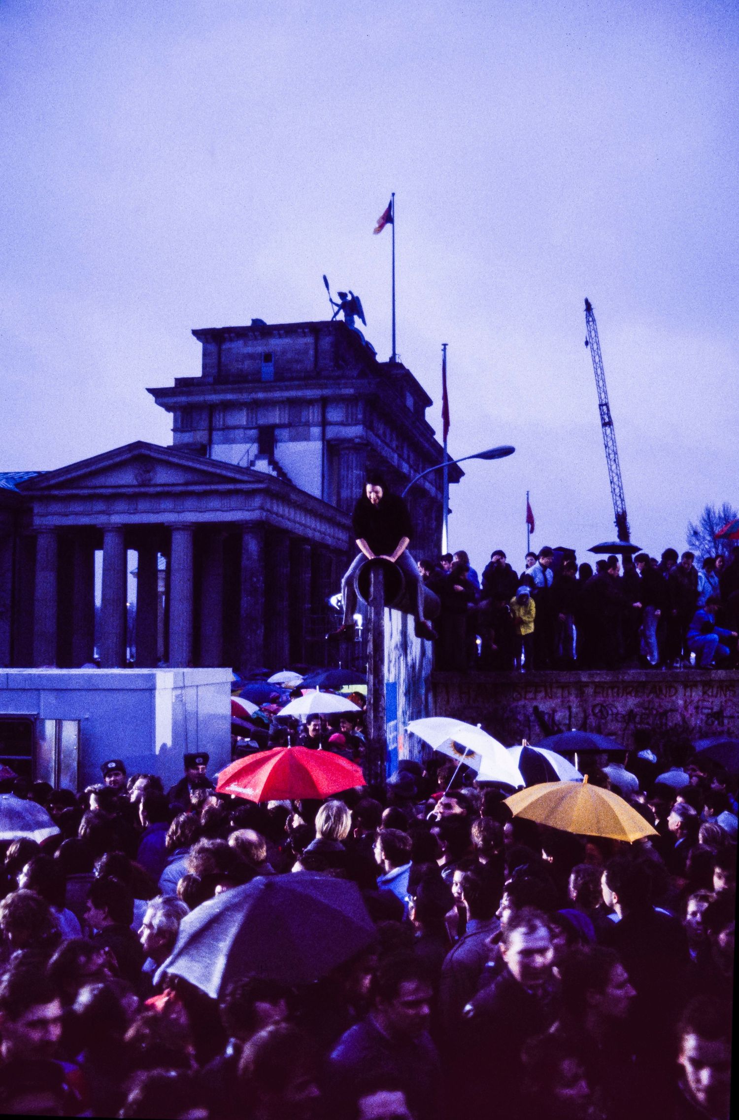 Large crowd gathered at Brandenburg Gate during twilight with umbrellas and flags visible.