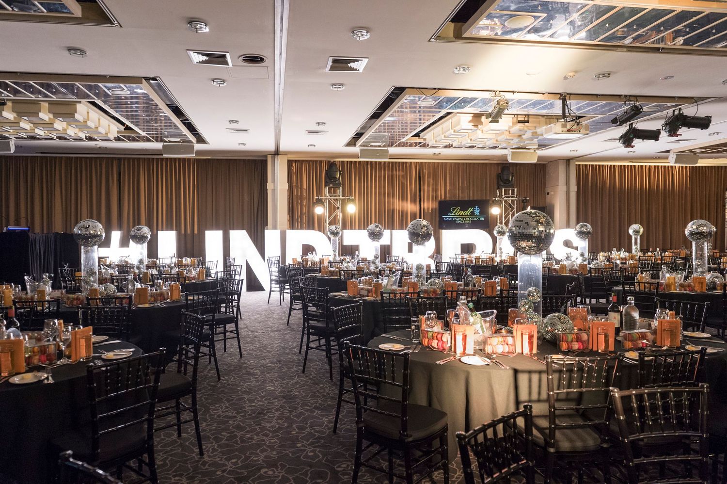Elegant banquet hall setup with round tables, dark chairs, and crystal chandeliers in a spacious event venue.