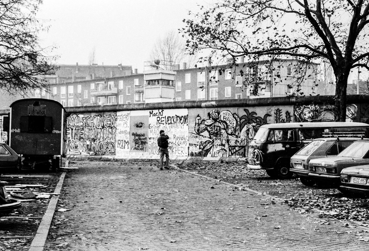 Berlin Wall segment covered in graffiti with parked vehicles and trees visible.