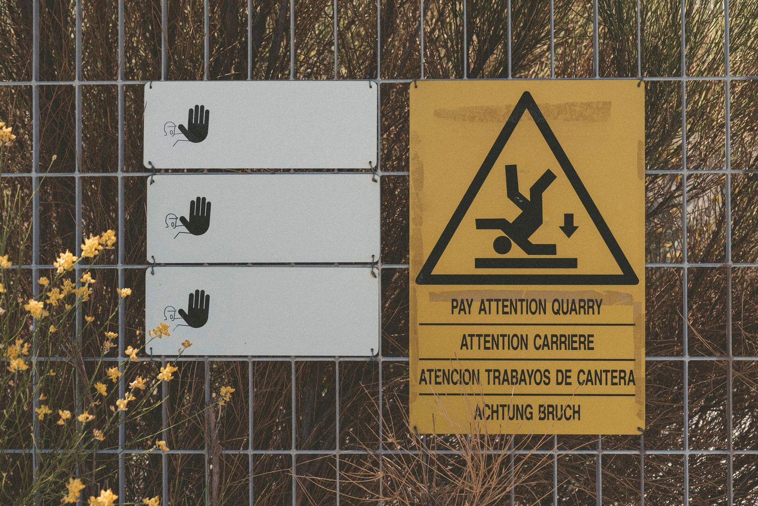 Warning sign showing falling hazard symbol with hand prints on white panels mounted on wire fence.