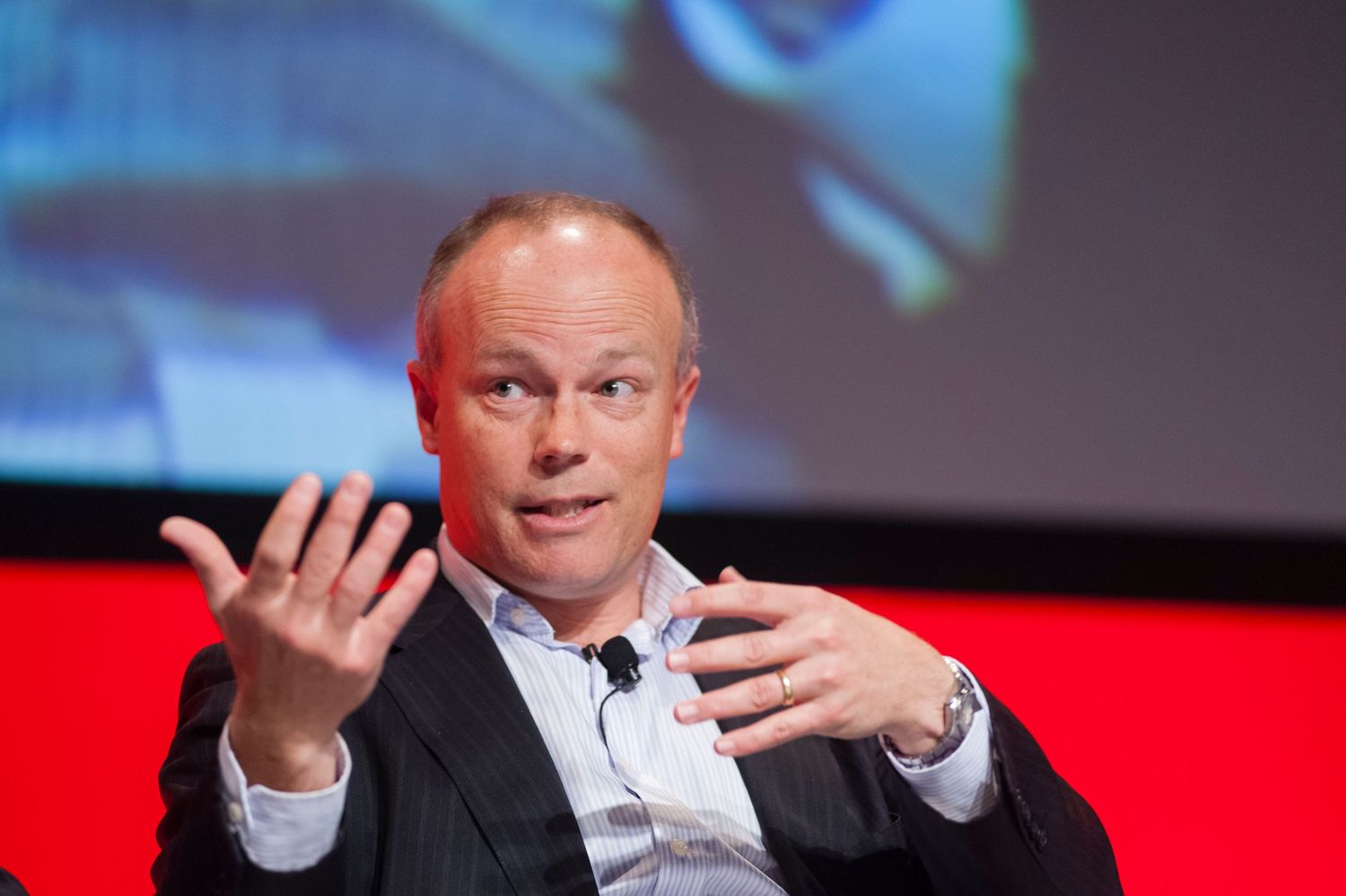 Speaker gesturing during presentation with vibrant digital displays in background.