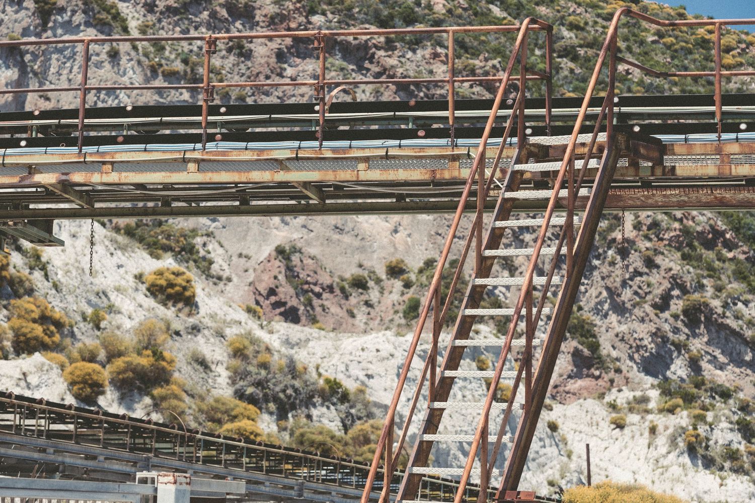 Metal ladders and walkways traverse rocky terrain at an industrial mining or construction site in mountainous landscape.