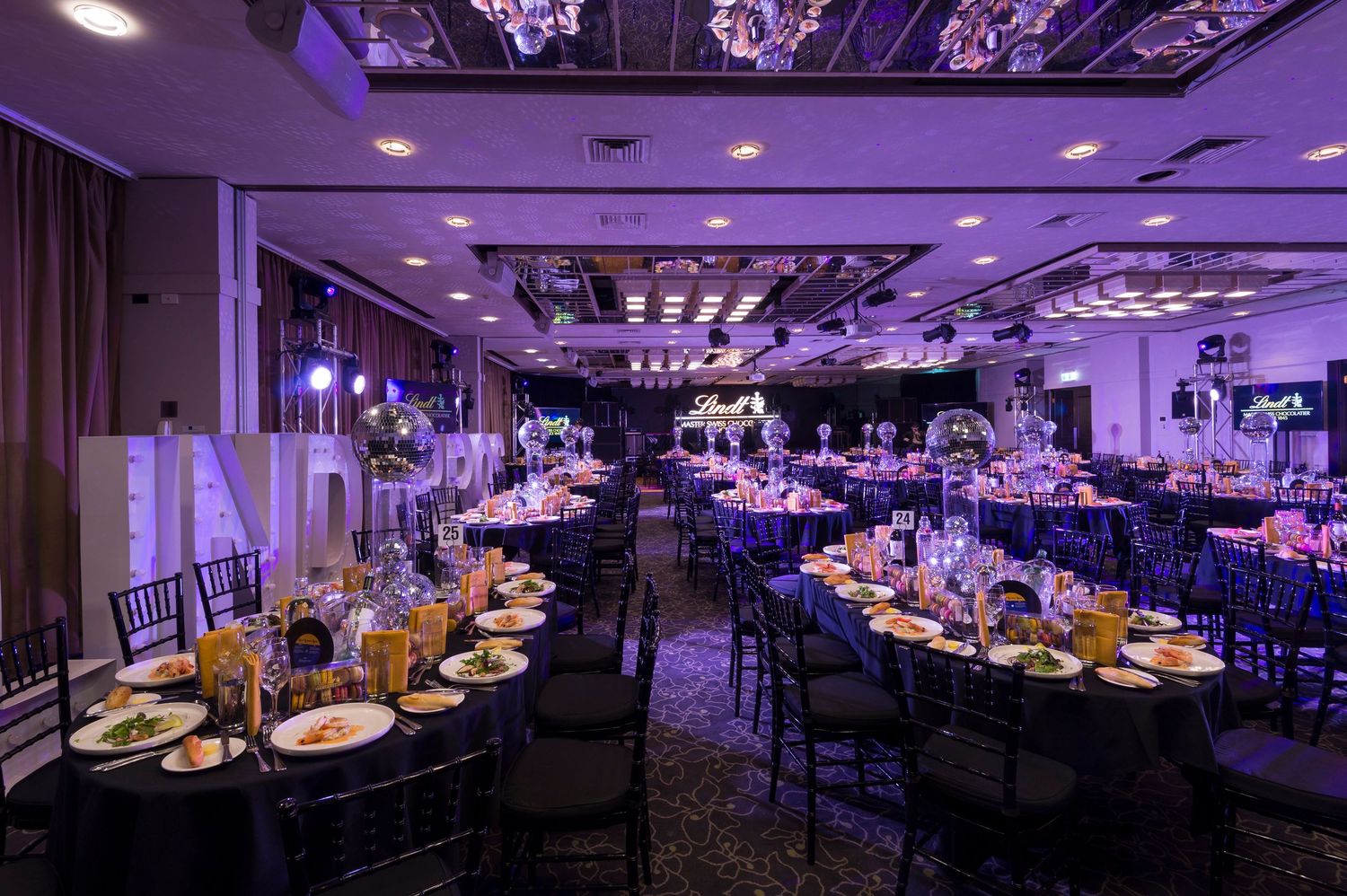 Elegant banquet hall with purple uplighting, crystal chandeliers, and round tables set for a formal dining event.