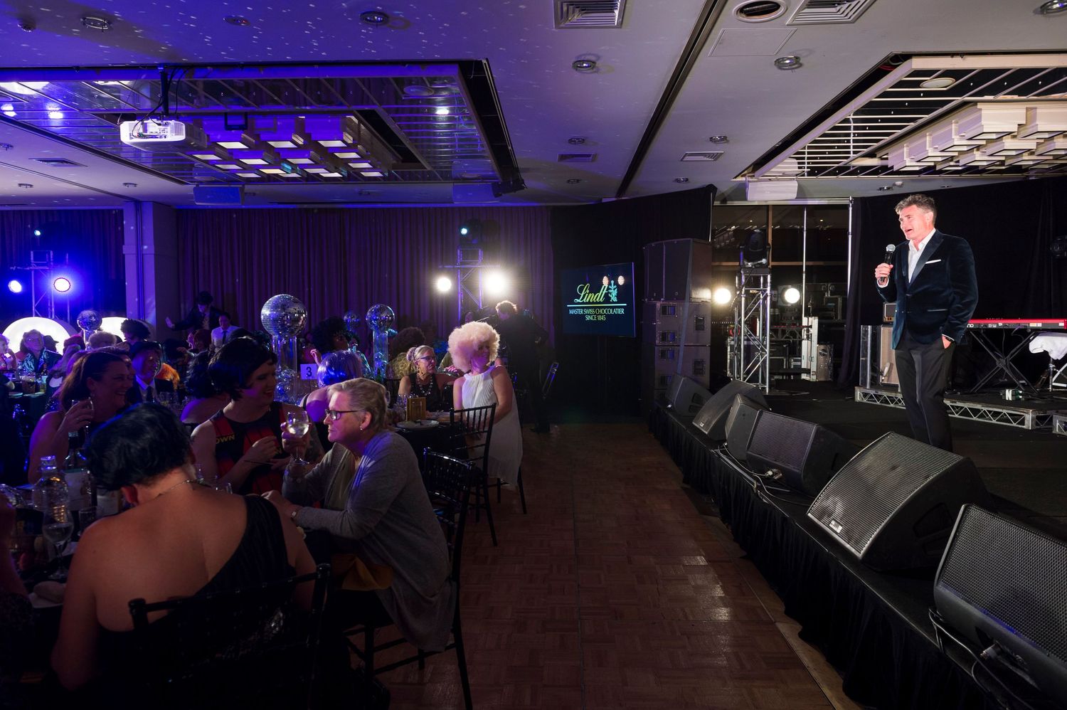 A performer speaks on stage in a dimly lit venue with blue lighting while audience members sit at tables.